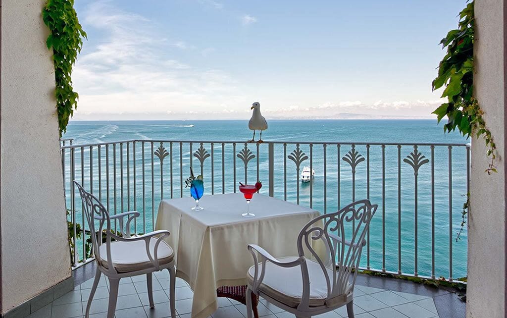 A balcony with a table and chairs overlooking the sea, featuring a seagull and a decorative bird sculpture.