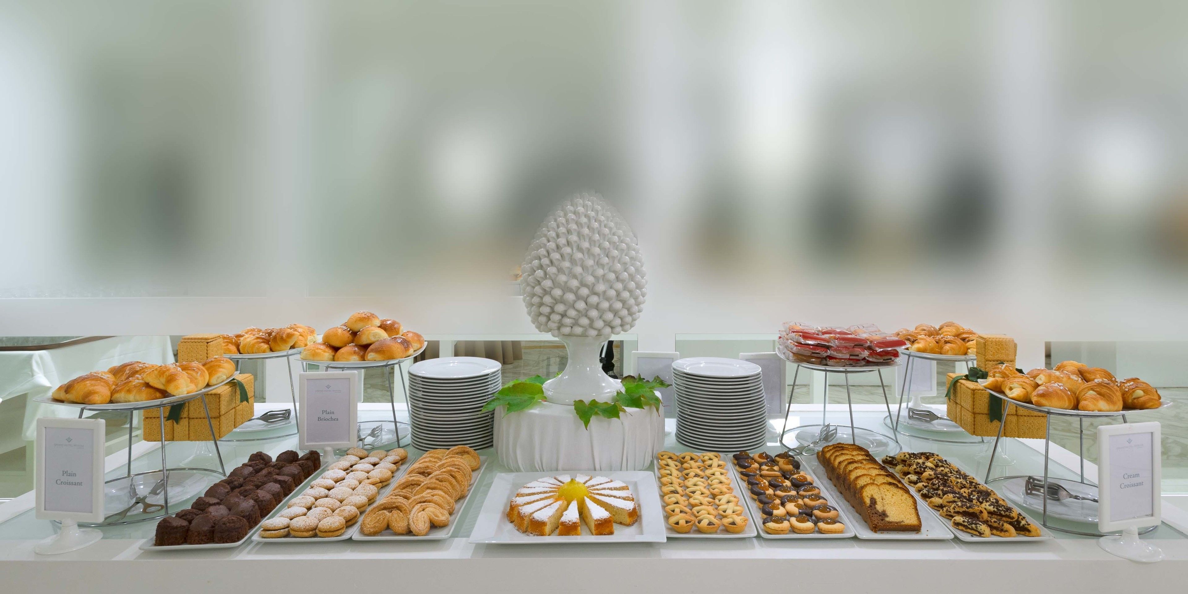 A buffet table with various pastries and desserts, including croissants, muffins, and a cake, arranged on plates and stands.