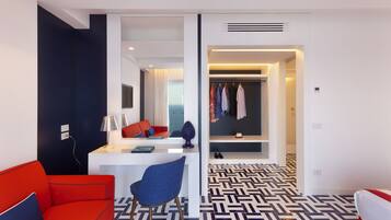 Une chambre d’hôtel moderne avec un canapé rouge, une chaise bleue, un bureau avec une lampe et un sol à motifs.