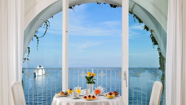 Une table à manger recouverte d’une nappe blanche, deux chaises et une vue sur la mer, visible depuis un balcon agrémenté de rideaux blancs et de plantes vertes.