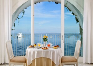 A dining table with a white tablecloth, two chairs, and a view of the sea through a balcony with white curtains and greenery. - Grand Hotel Riviera (Sorrento)