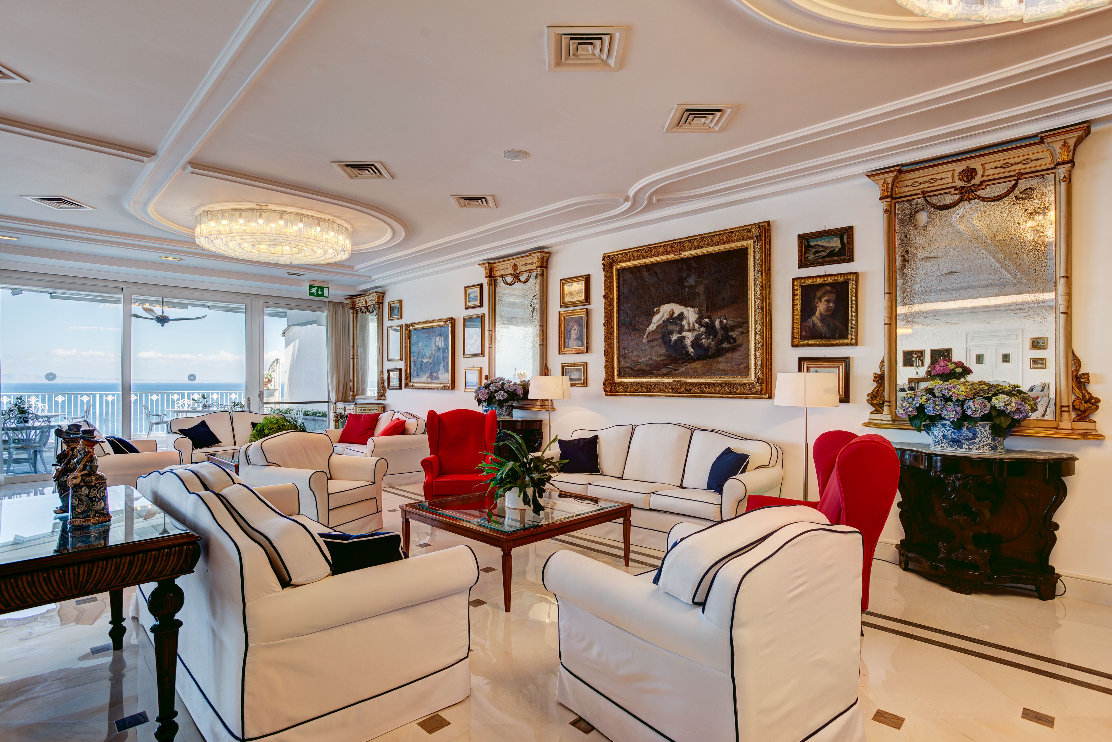 A spacious, elegantly furnished lounge with a large window offering a city view, a chandelier, and a variety of framed artworks on the walls.