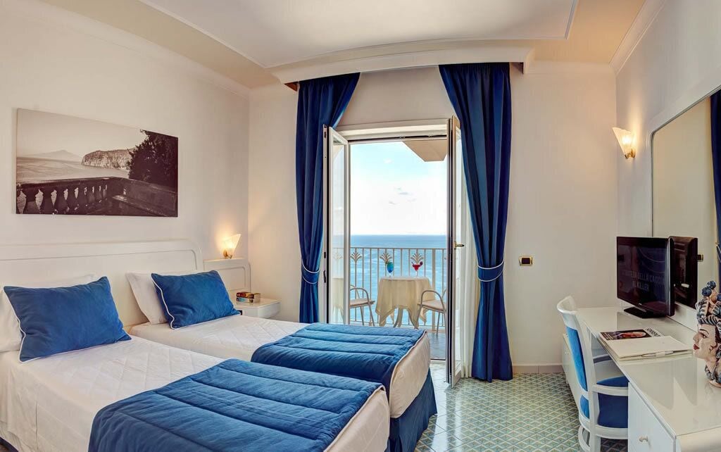 A hotel room with two beds, a desk, a TV, and a balcony with a table and chairs.