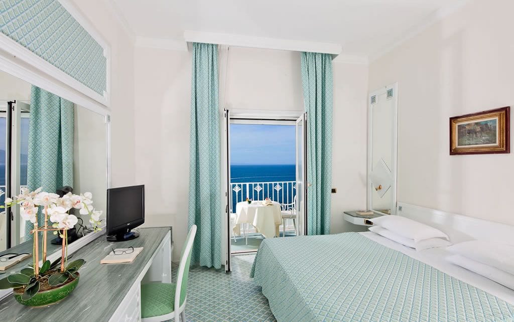 A hotel room with a bed, a desk with a computer, a chair, a small table with a plant, and a view of the sea through a balcony door.