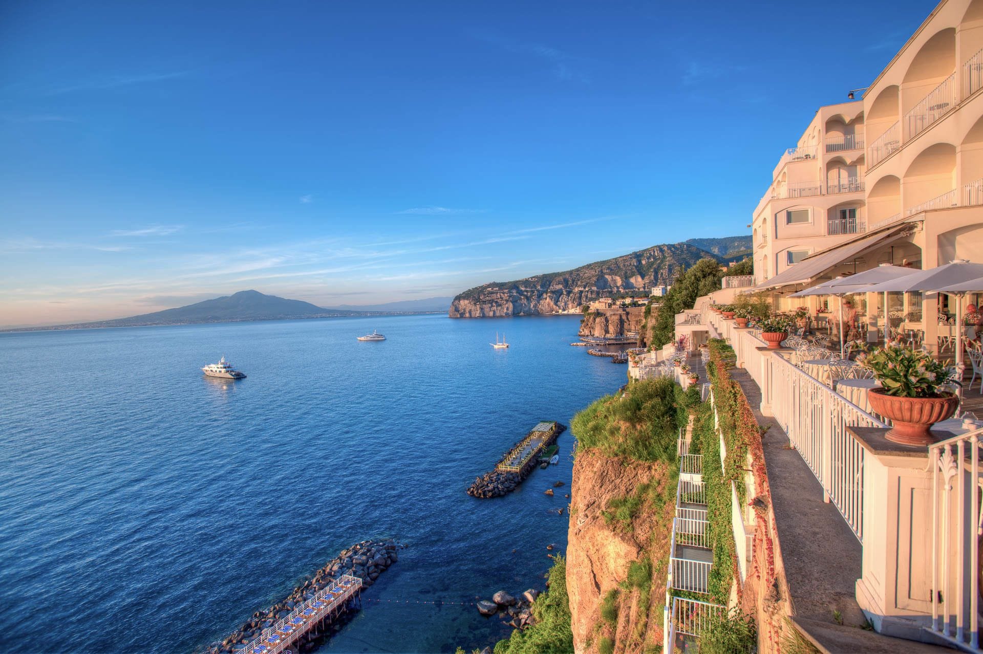 Un hotel costero con un balcón alargado con vistas al mar, un muelle que se adentra en el agua y montañas al fondo.