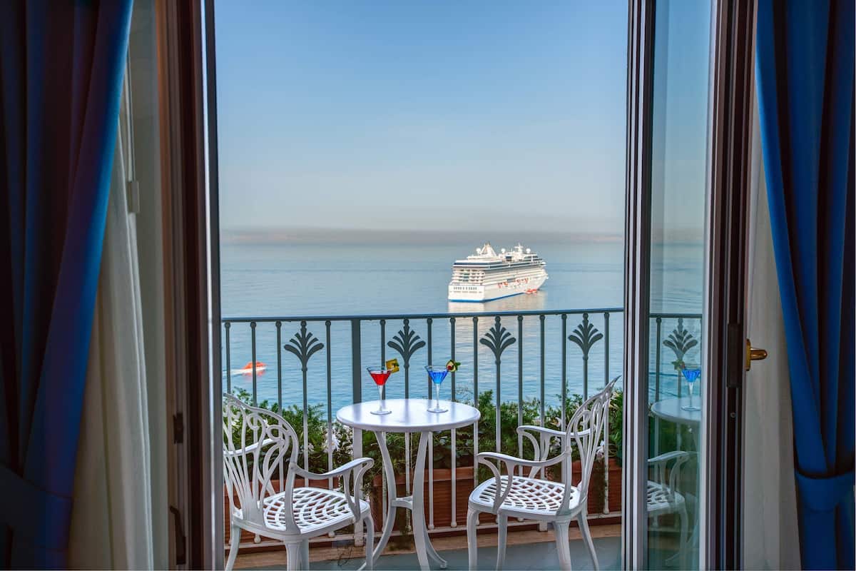 Ein Balkon mit weißen Stühlen und einem Tisch, mit Blick auf ein ruhiges Meer, in der Ferne ein Kreuzfahrtschiff.