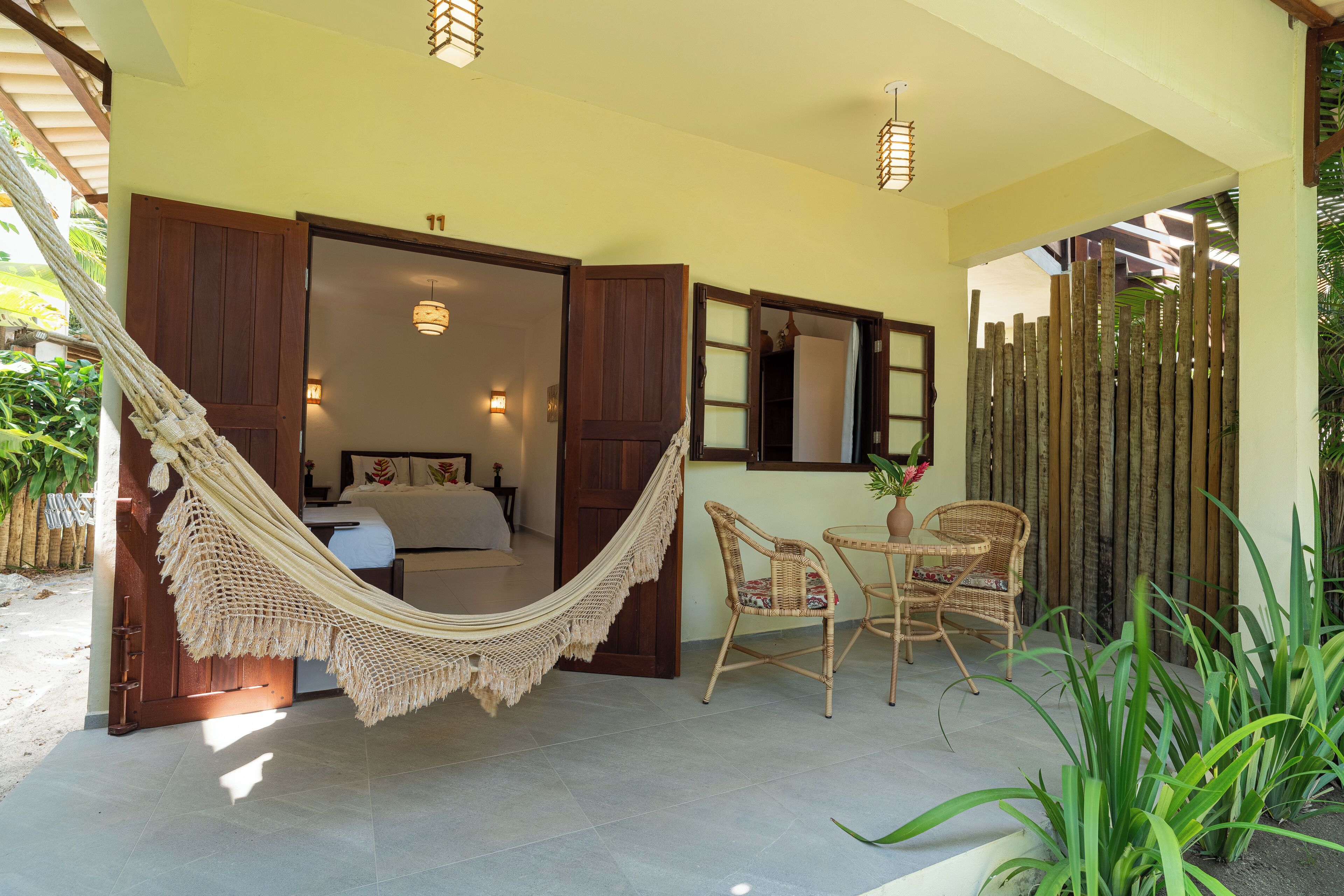 A room with a hammock, wicker chairs, a small table, and a bed with a view of the garden.