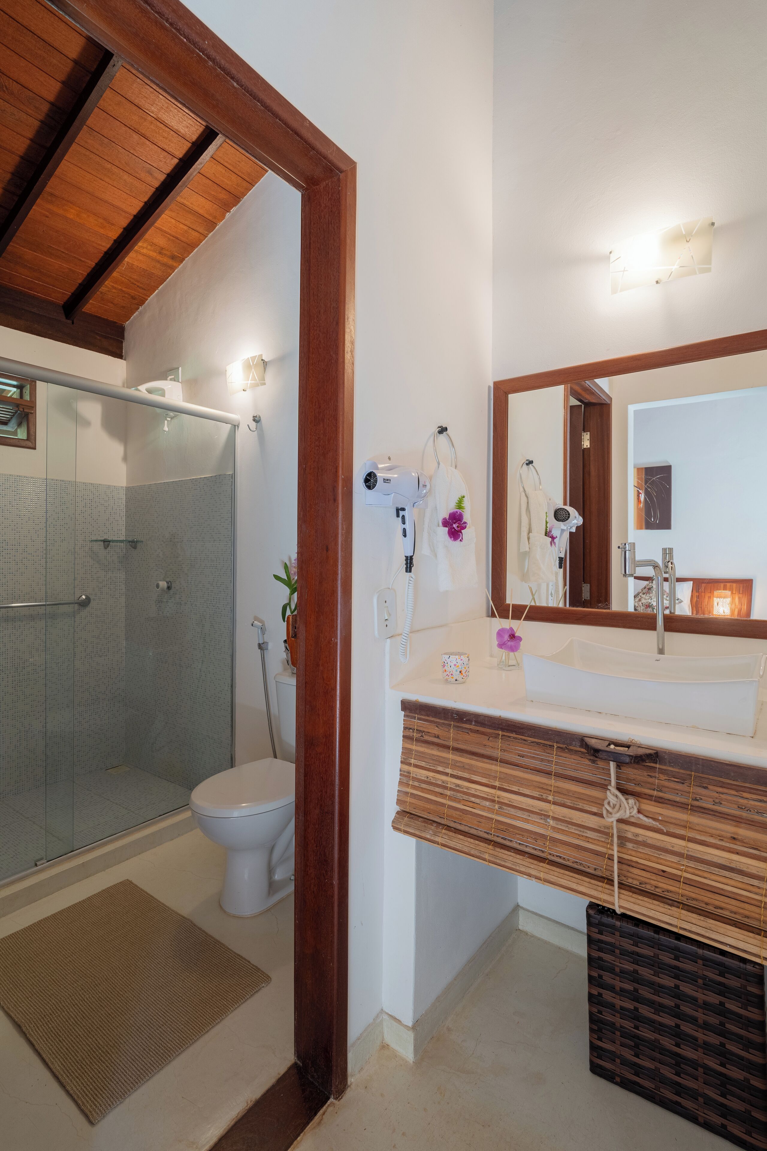 A bathroom with a glass shower enclosure, a white toilet, a wooden vanity with a mirror, and a small plant on the counter.