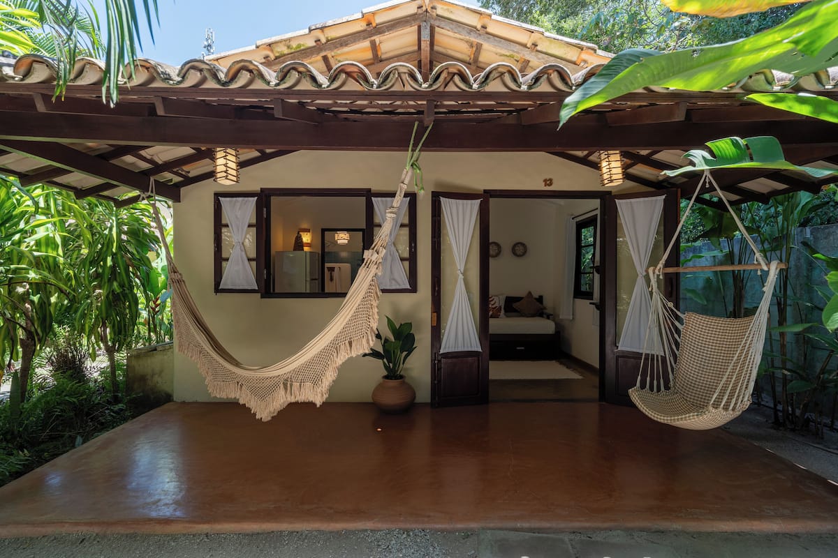 A covered outdoor area with a hammock, a potted plant, and a view into a room with a bed and curtains.
