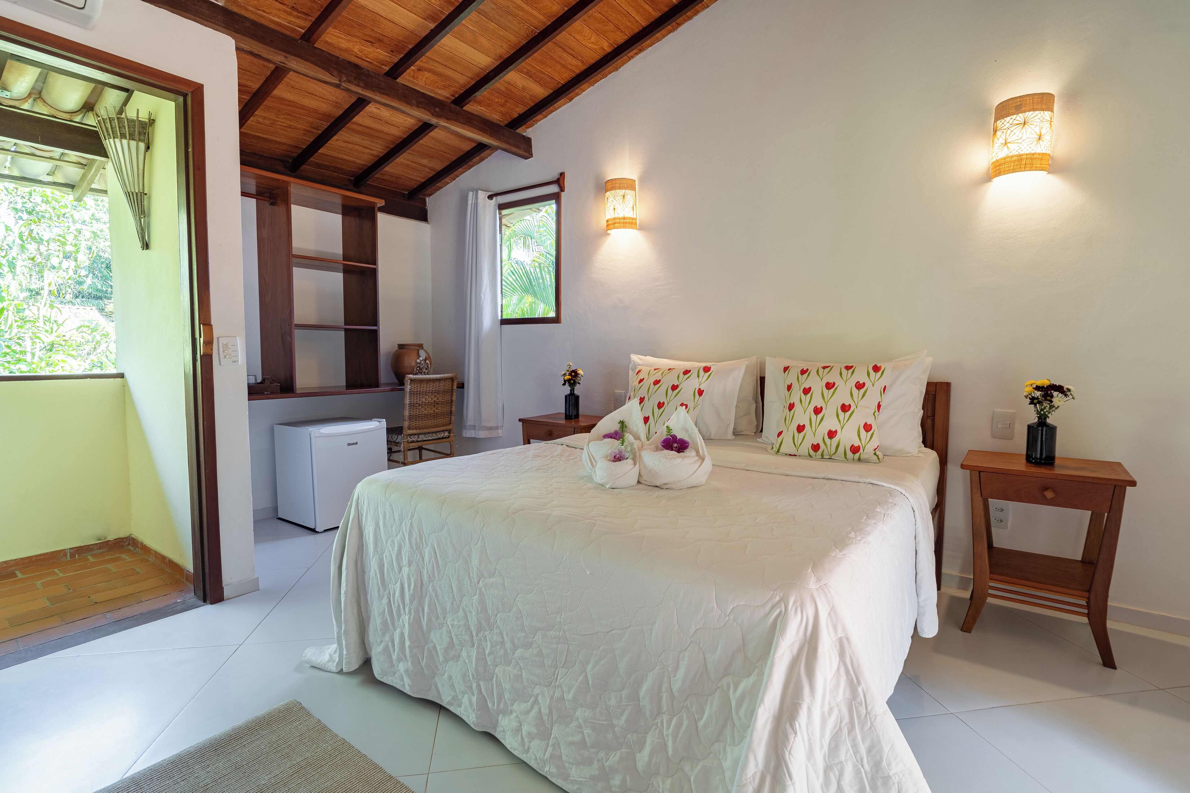 A bedroom with a large bed, two bedside tables, a nightstand, a window with curtains, a small desk, and a refrigerator.