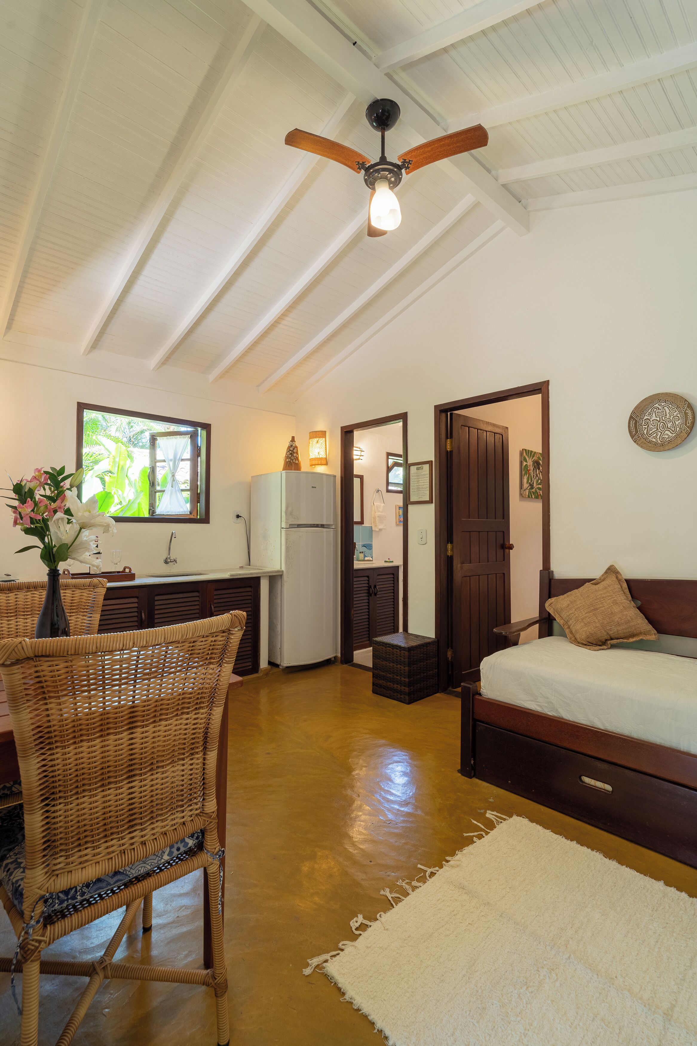 A room with a bed, a dining table with chairs, a ceiling fan, a refrigerator, and a window with a view of greenery.