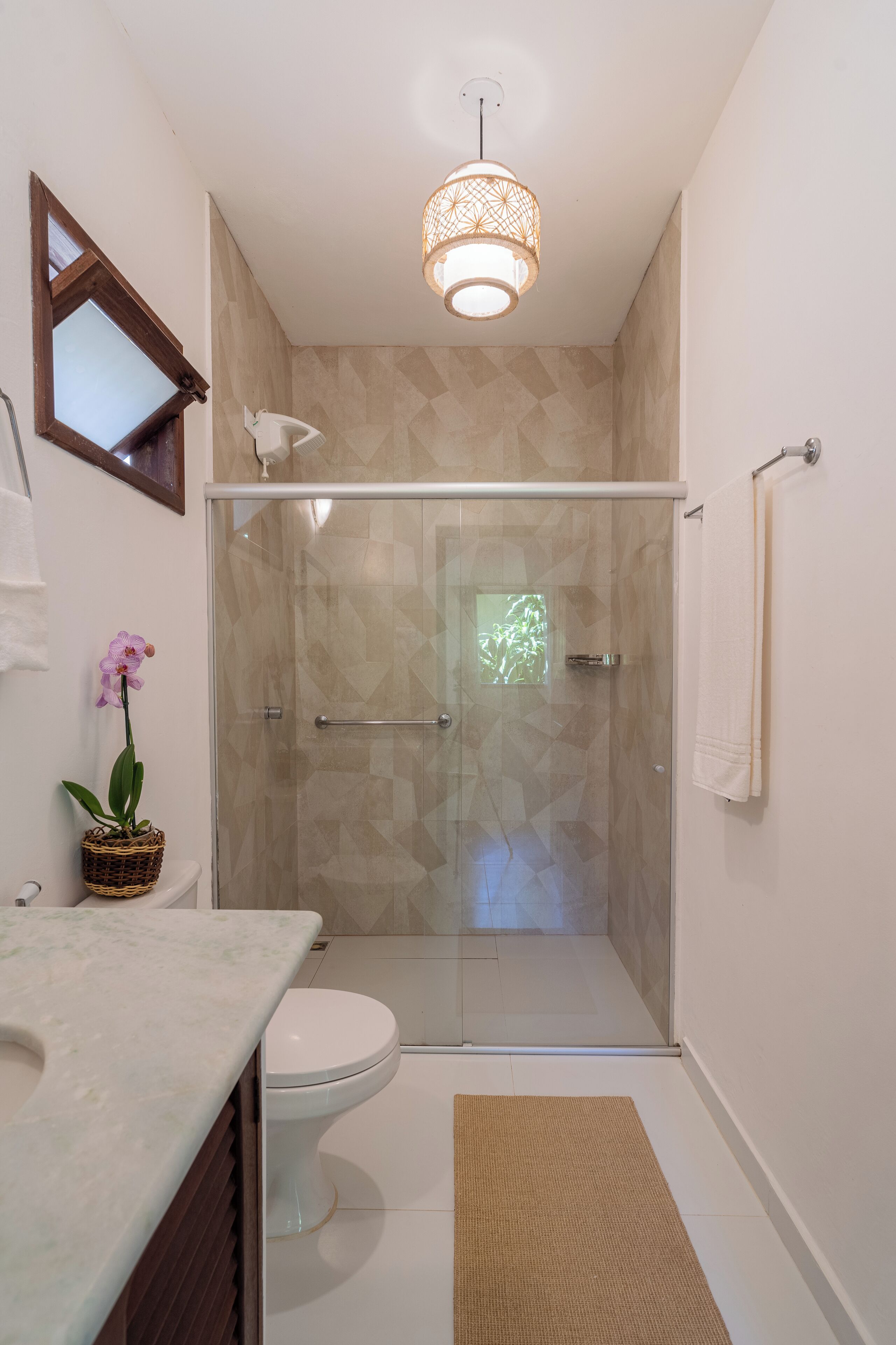 Ein Badezimmer mit einer ein Glasfach raumseitig abgetrennten Dusche, einer weißen Toilette, einem Marmorwaschbecken, einem Korbwäscher mit einer Orchidee, einer Pendelleuchte und einem Handtuchhalter.