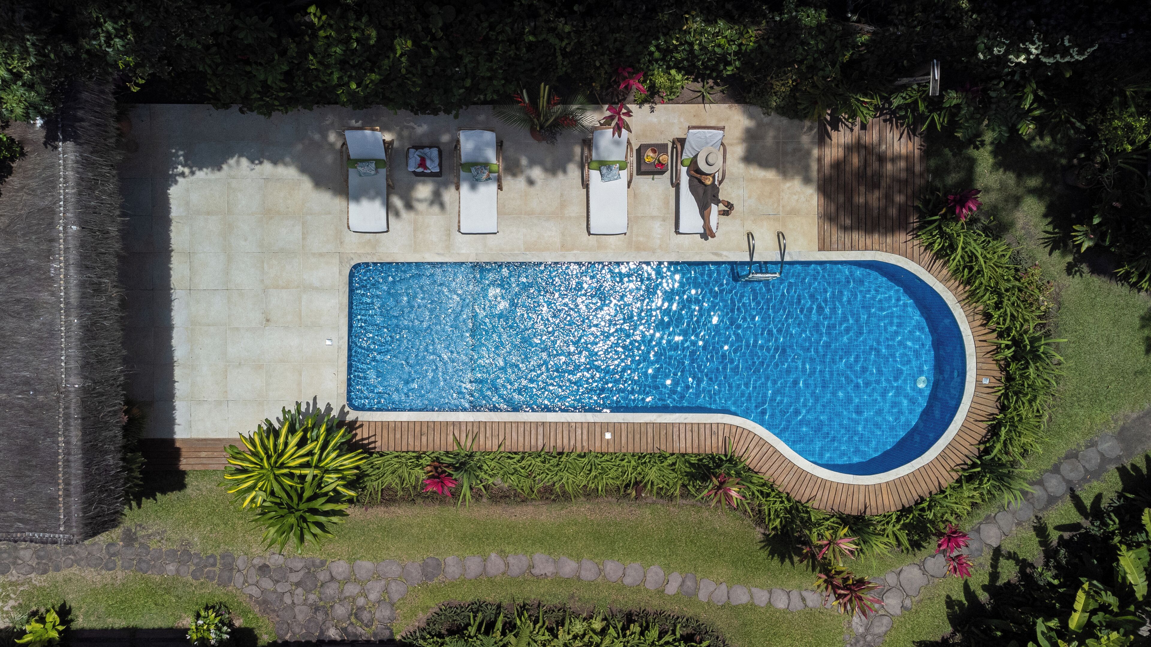 An aerial view of a rectangular swimming pool with a wooden deck surrounding it, accompanied by lounge chairs and a small table with a towel.