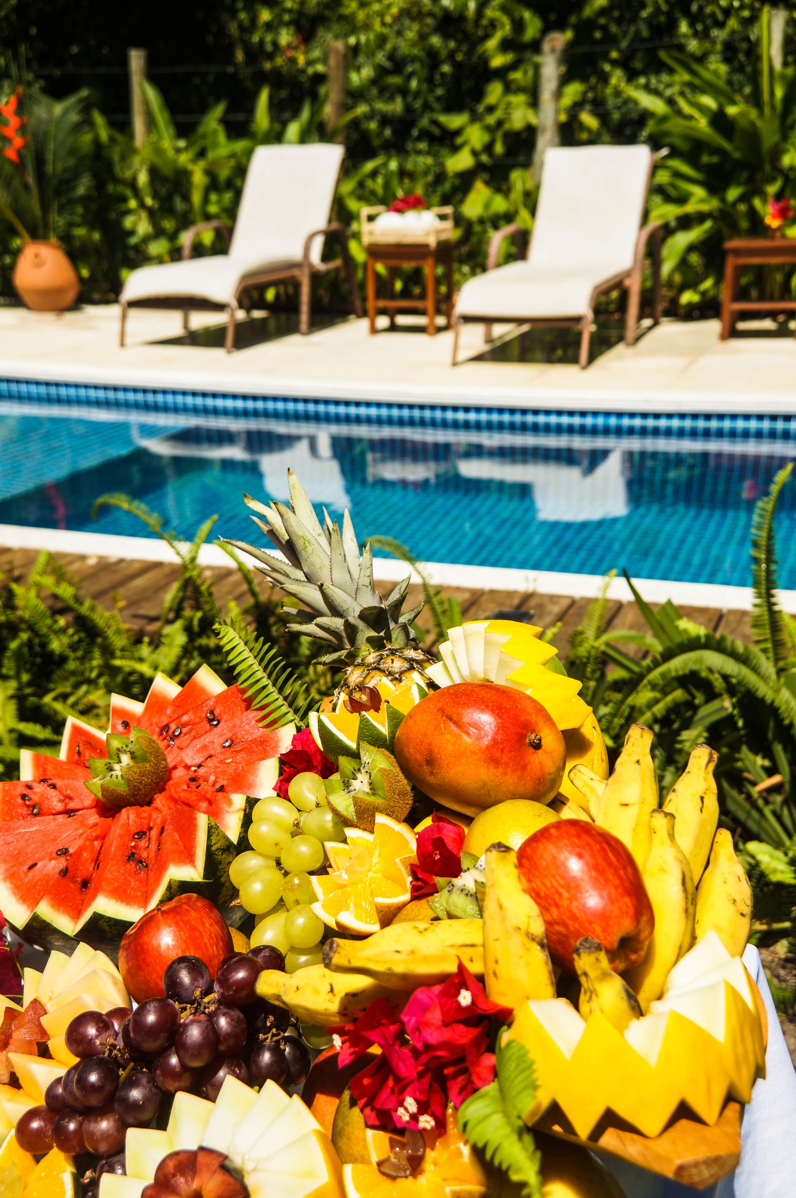Une présentation de fruits au bord de la piscine, avec la piscine et des chaises longues en arrière-plan.