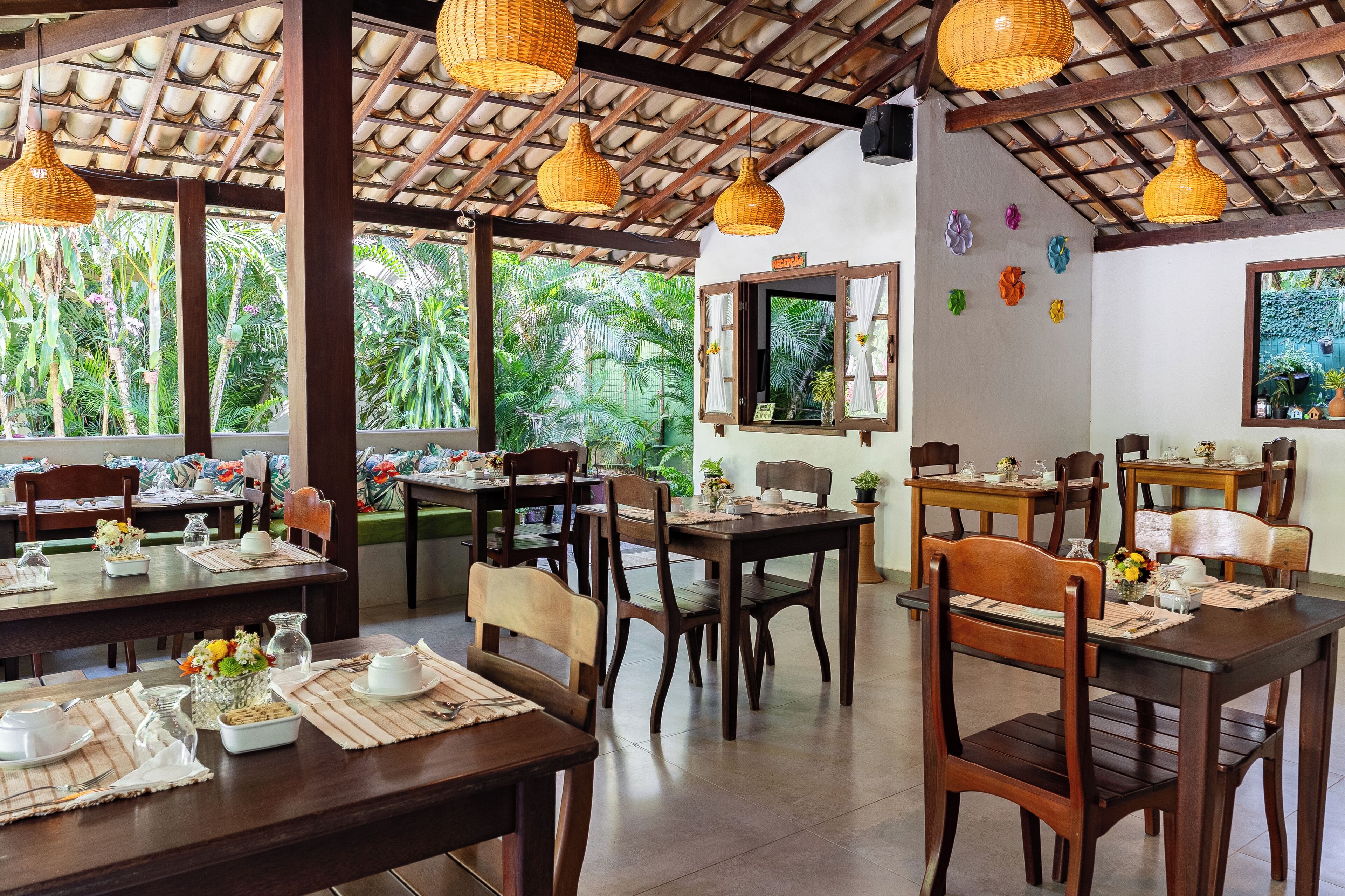 A restaurant with wooden tables and chairs, hanging wicker lamps, and a view of lush greenery outside.