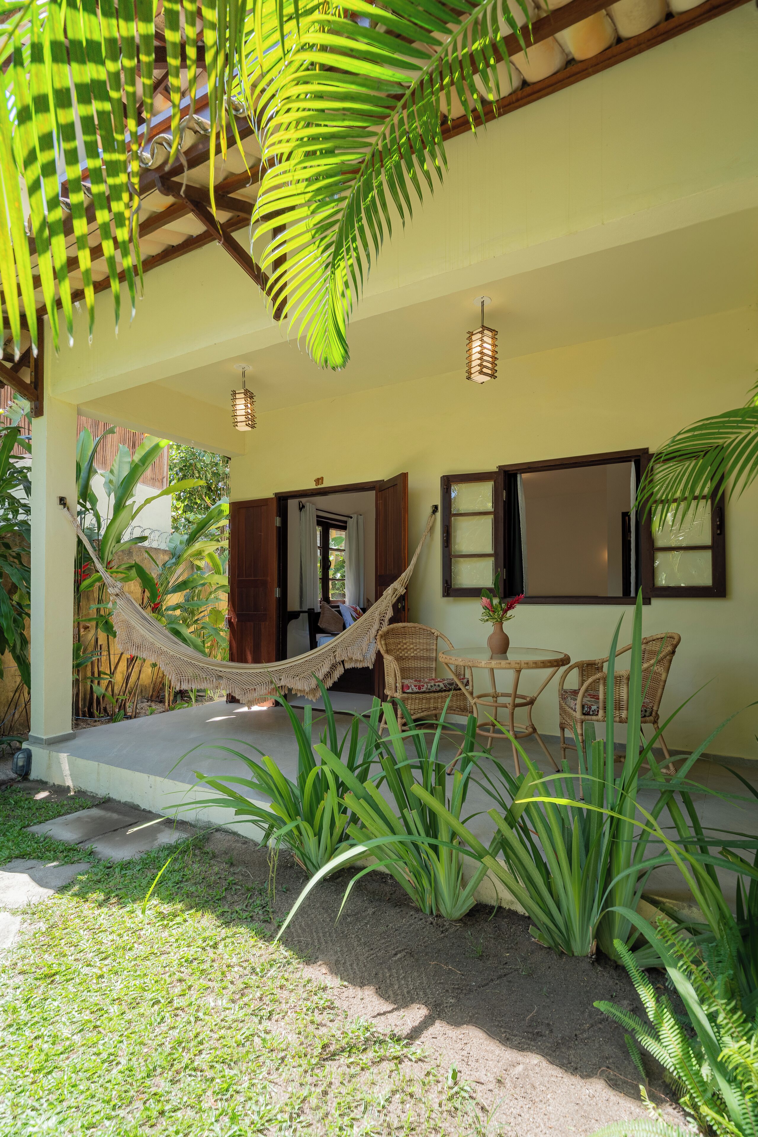 A covered outdoor seating area with wicker furniture, a hammock, and lush greenery.
