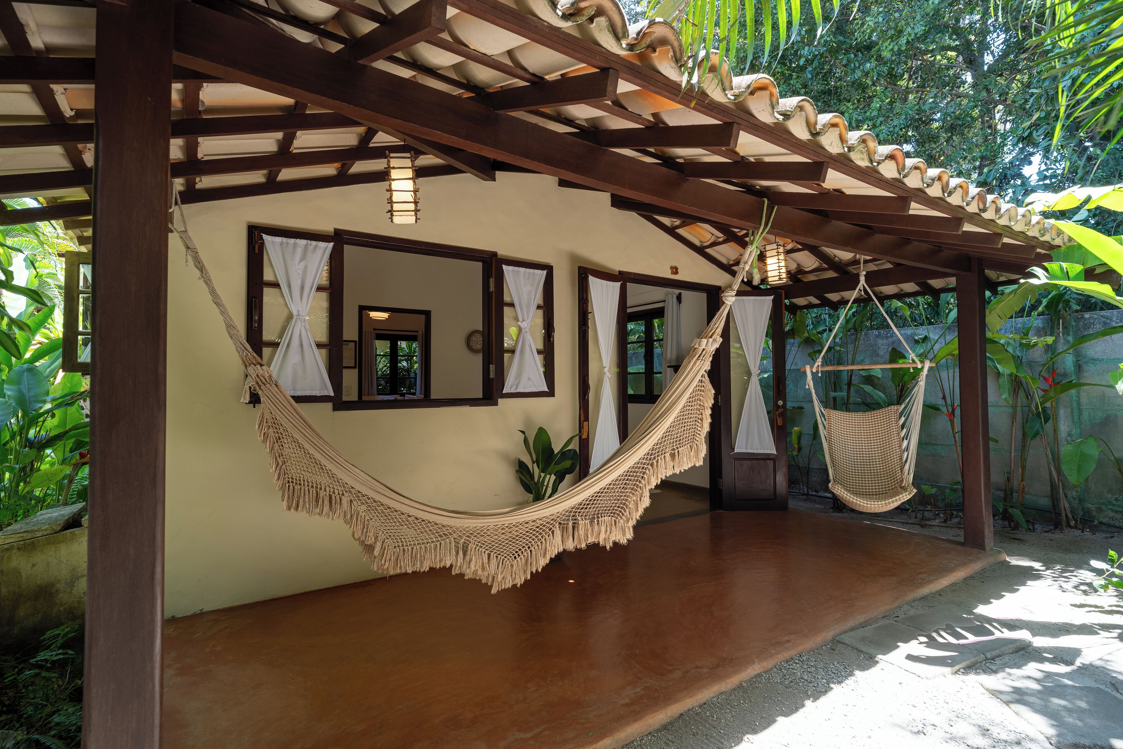 A hammock under a wooden pergola, a small plant, and a view of a lush garden.
