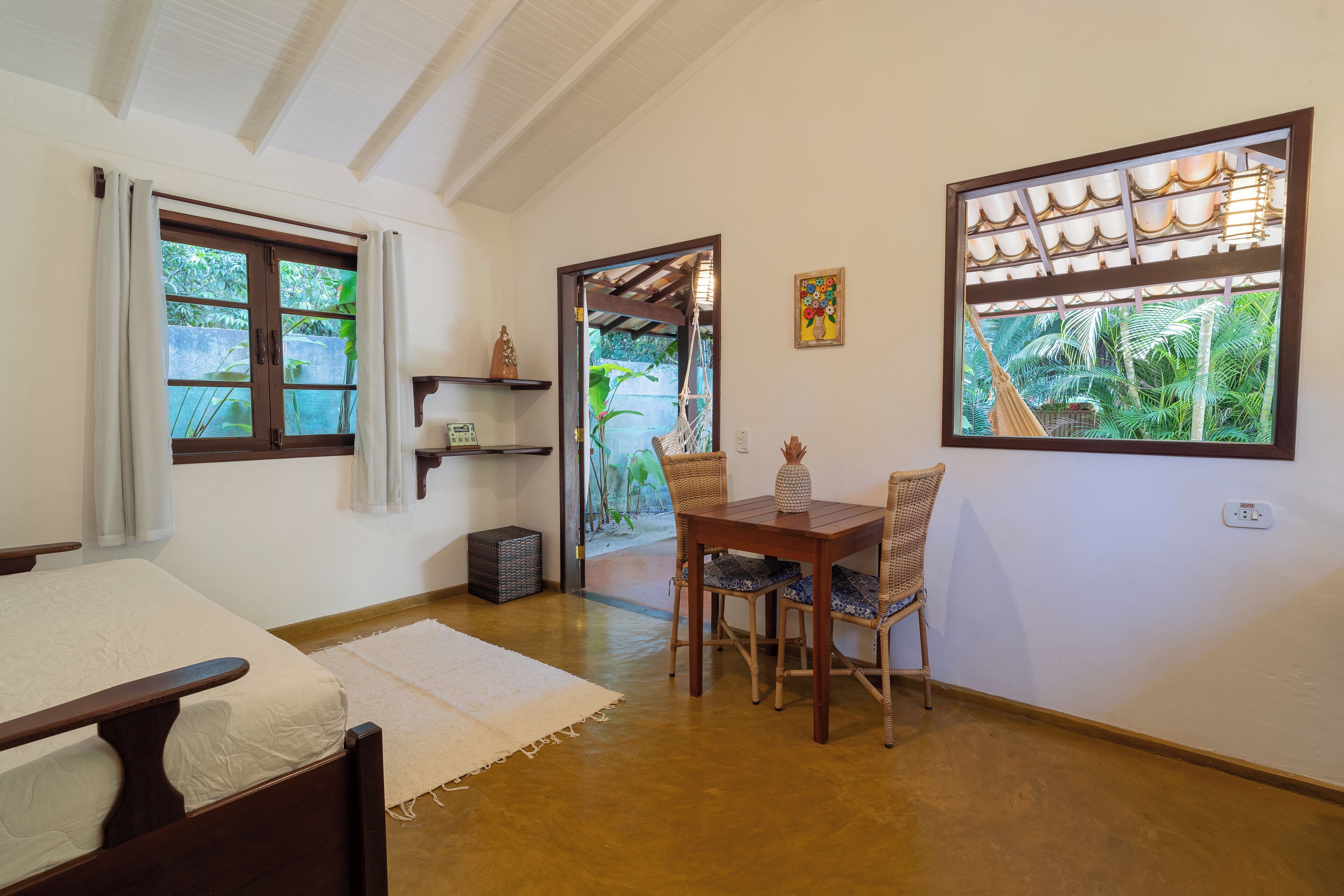 A room with a bed, a dining table with chairs, a window with a view of greenery, a door leading outside, and a shelf with decorative items.