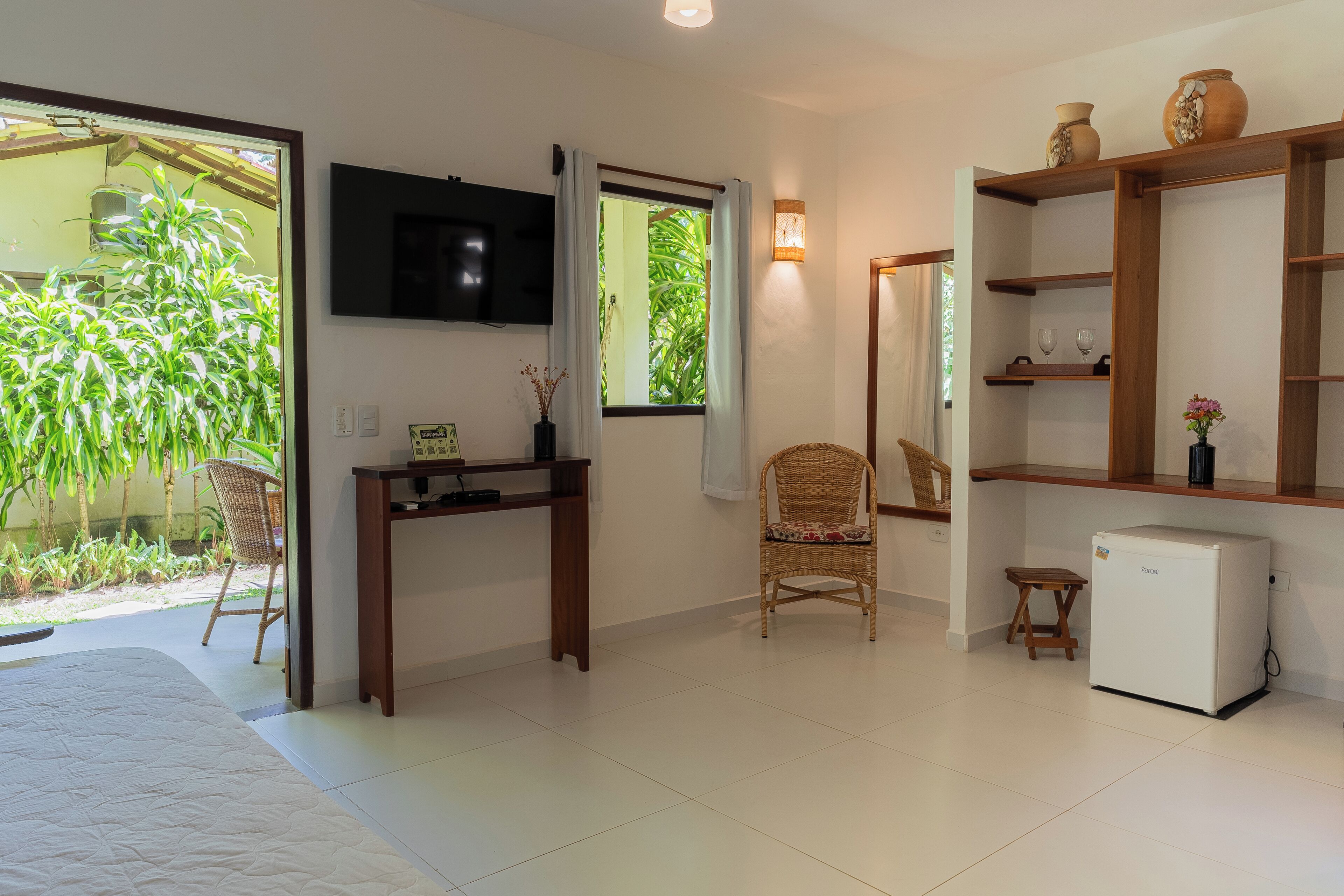 A bedroom with a bed, a TV, a desk, a chair, a small refrigerator, and a window with a view of greenery.