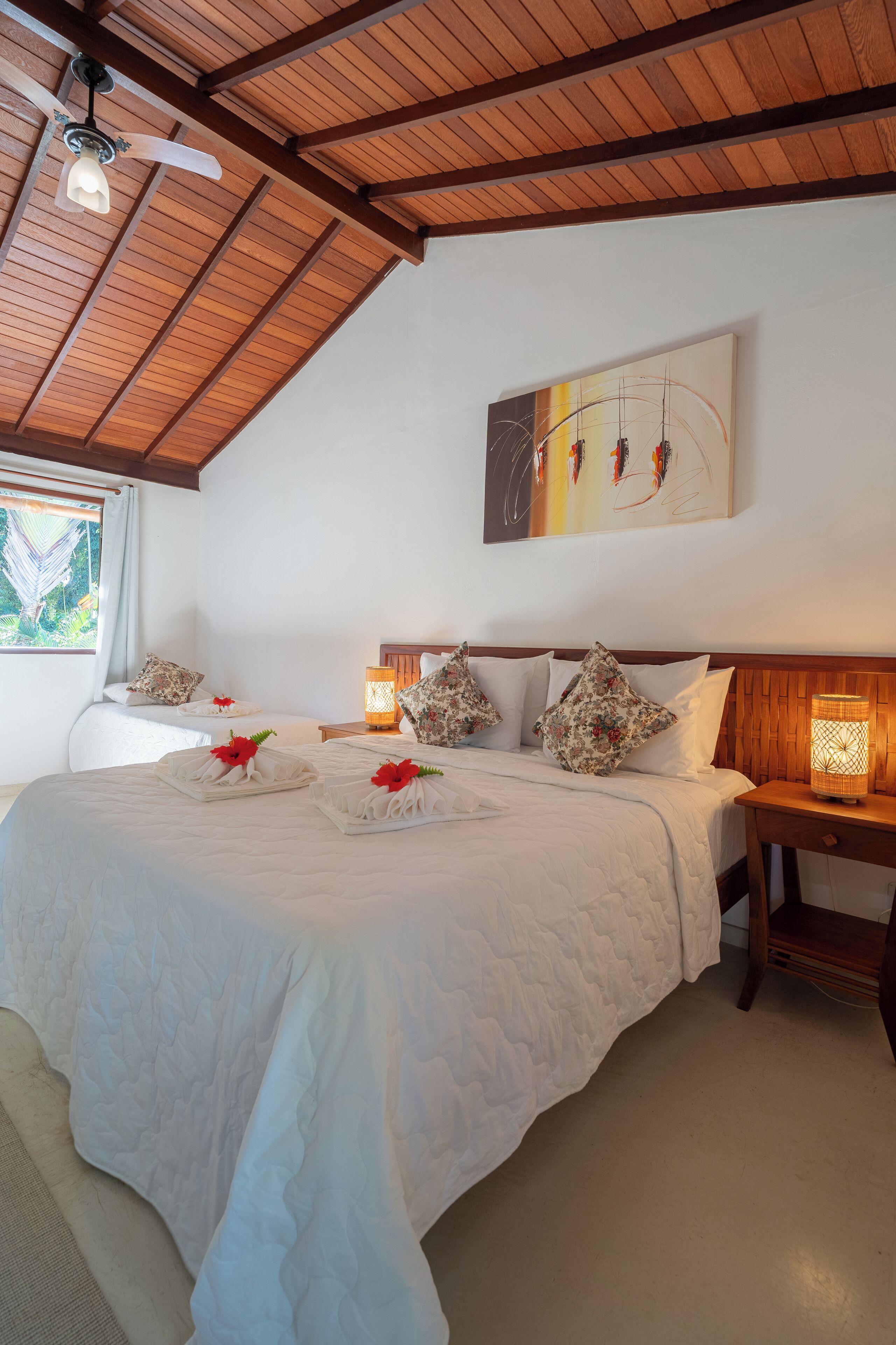 A hotel room with a wooden ceiling, a large bed with white bedding, a nightstand with a lamp, and a painting on the wall.
