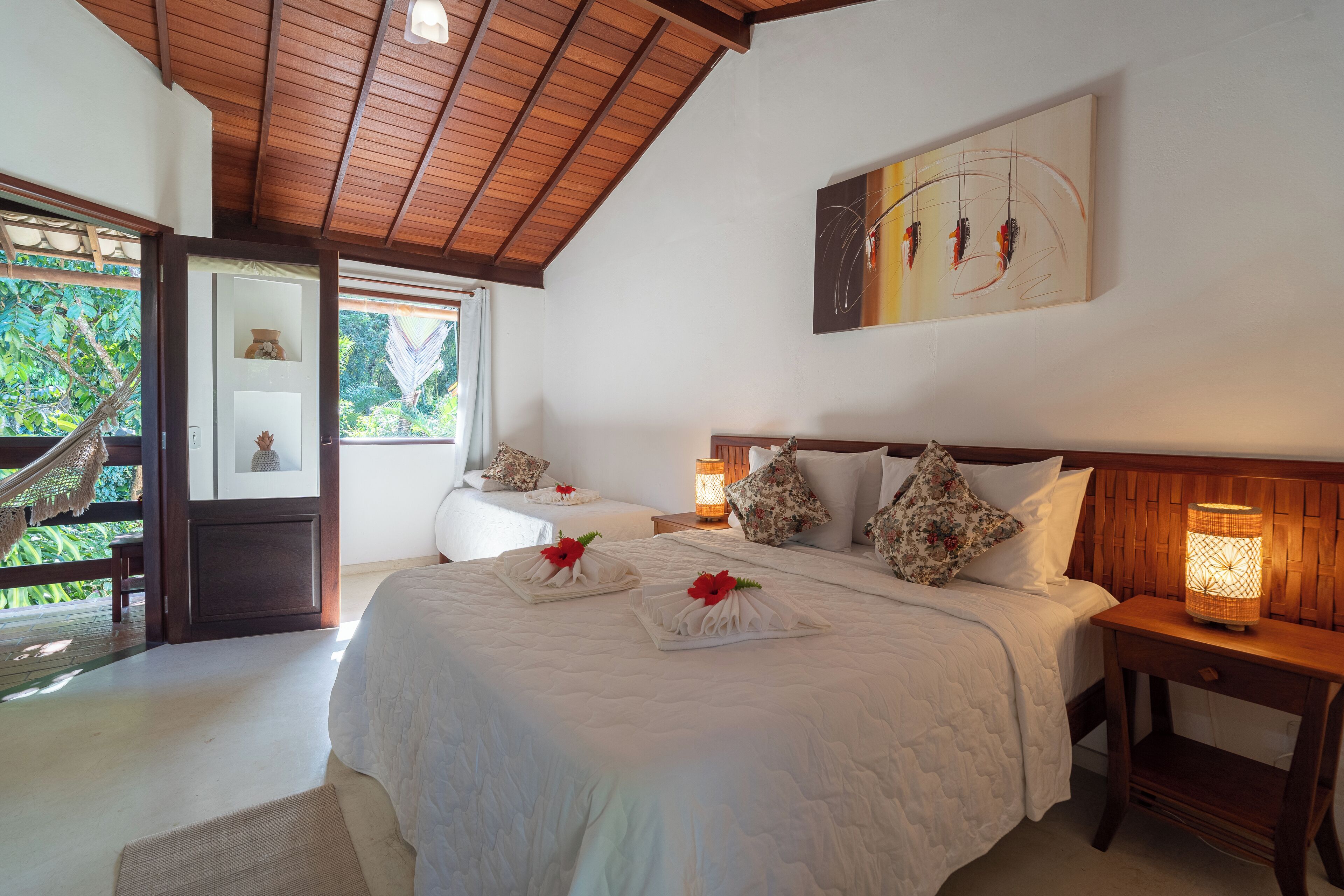 A bedroom with a large bed, a wooden headboard, a nightstand with a lamp, and a view of the outdoors.