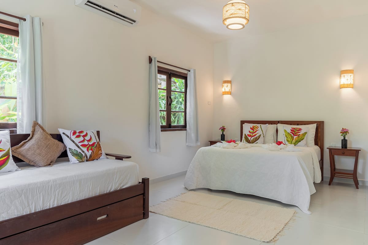 A bedroom with two beds, a large window with curtains, a ceiling fan, and wall-mounted lights.