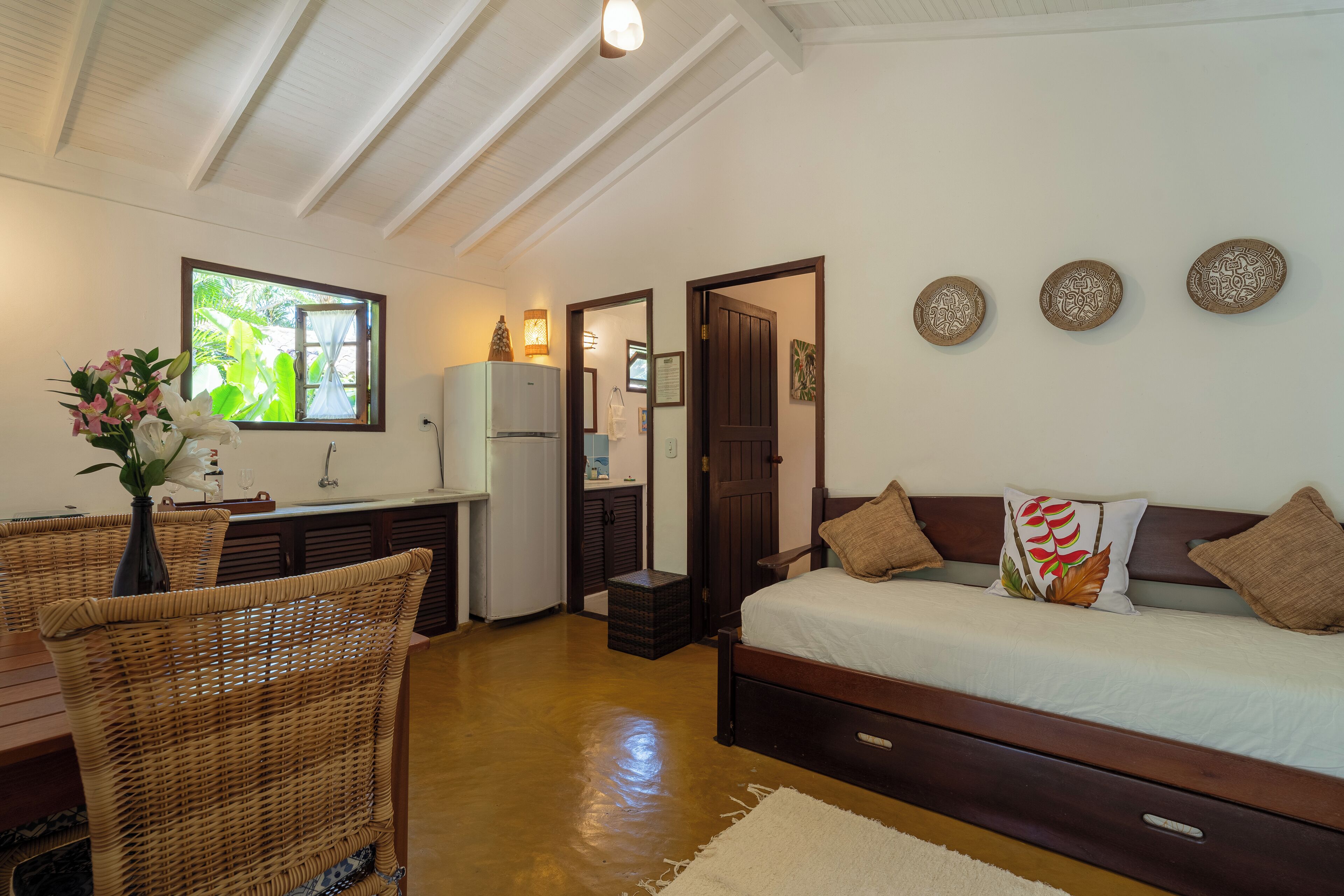 A bedroom with a bed, a nightstand, a dresser, a window, and a dining area with a chair and table.