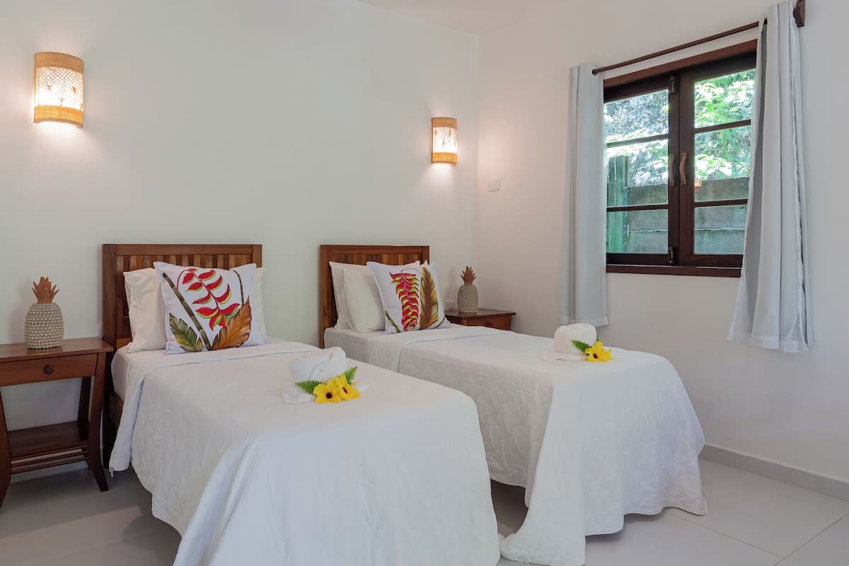 A hotel room with two beds, a wooden headboard, a nightstand, a lamp, and a window with curtains.