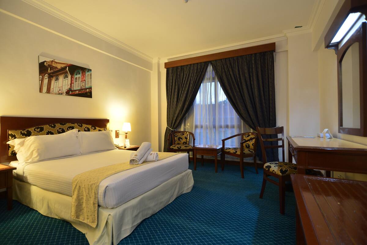 A hotel room with a bed, two chairs, a desk, a mirror, a painting on the wall, and a window with curtains.