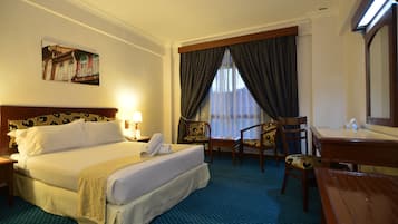 A hotel room with a bed, two chairs, a desk, a mirror, a painting on the wall, and a window with curtains.