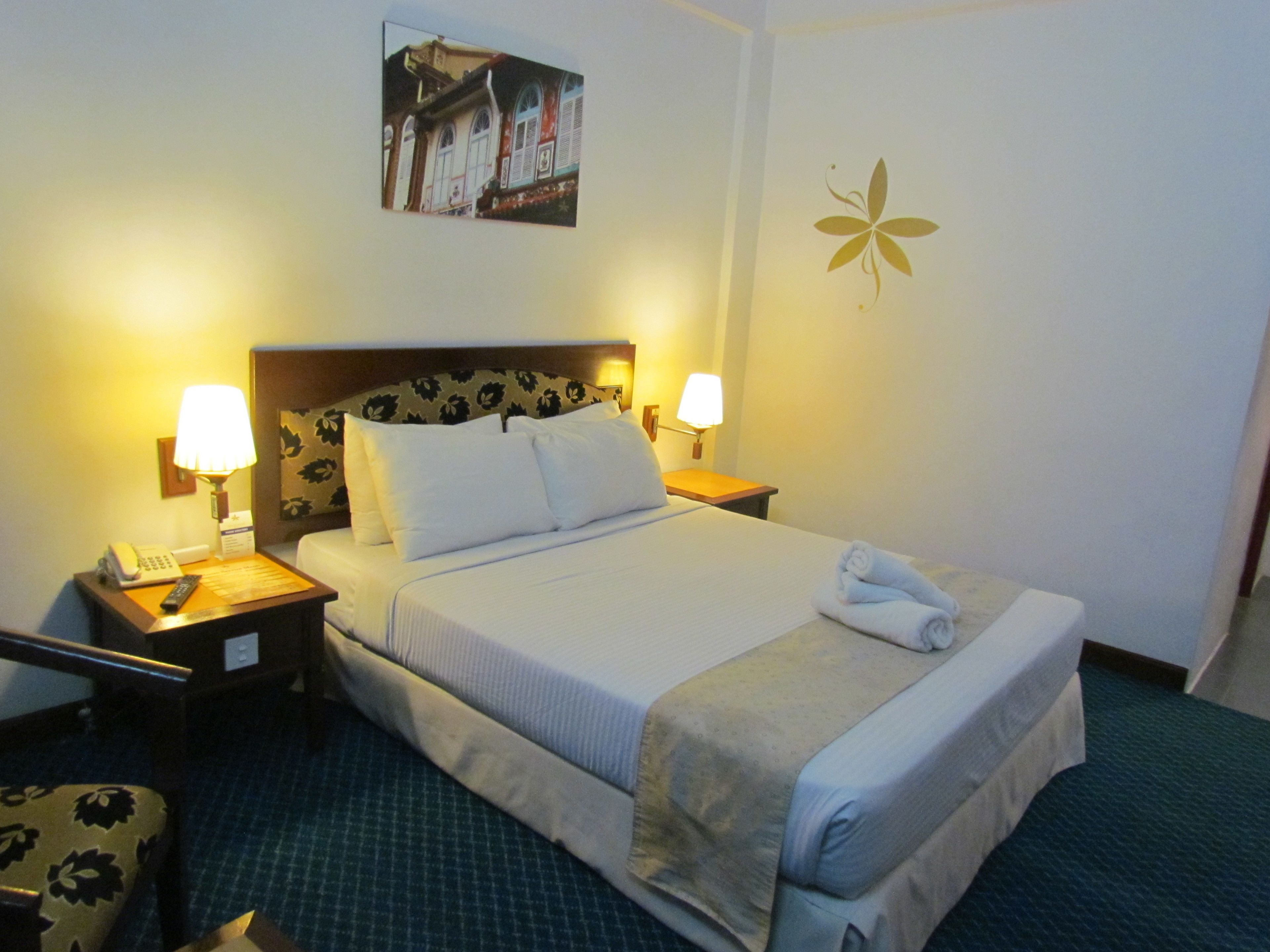 Habitación de hotel con cama, dos lámparas de noche, teléfono, una silla y un adorno en la pared.