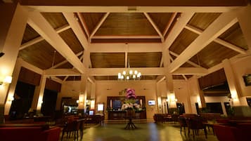 En rummelig lobby med høje lofter, træbjælker og en stor lysekrone i midten. Der er flere siddeområder med røde polstrede stole og en reception i baggrunden.