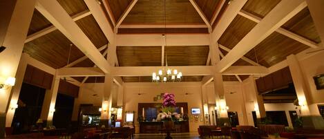 A spacious lobby with high ceilings, wooden beams, and a central chandelier. There are multiple seating areas with red upholstered chairs and a reception desk in the background.