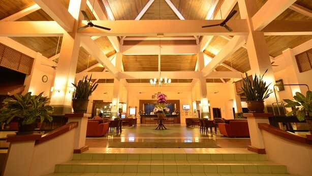 Een ruime lobby met een hoge plafond, houten balken en plafondventilatoren. Er staan verschillende potplanten en een receptiebalie in het midden.