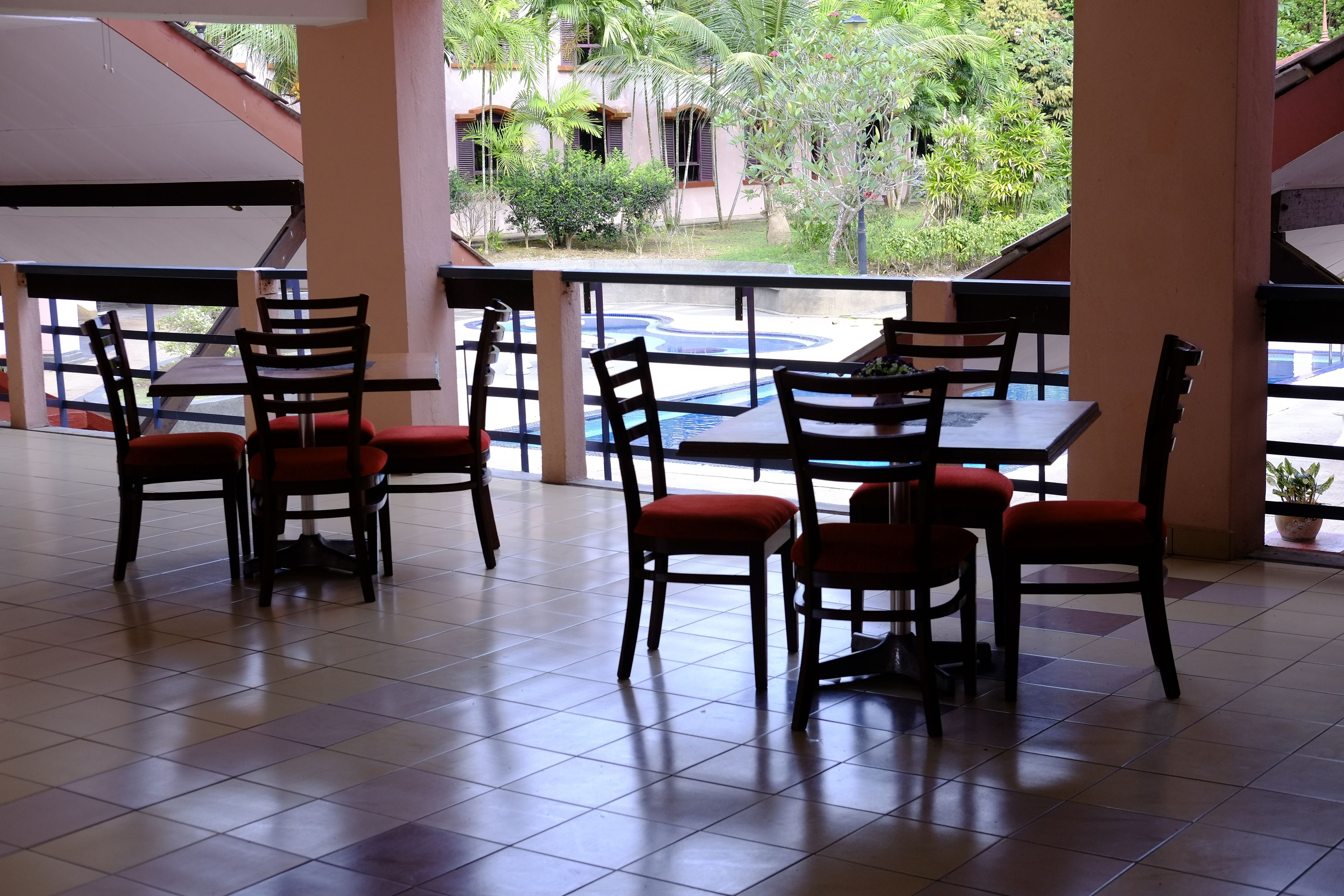 Un restaurante con terraza, que cuenta con mesas y sillas con cojines rojos, y vistas a una piscina y follaje.