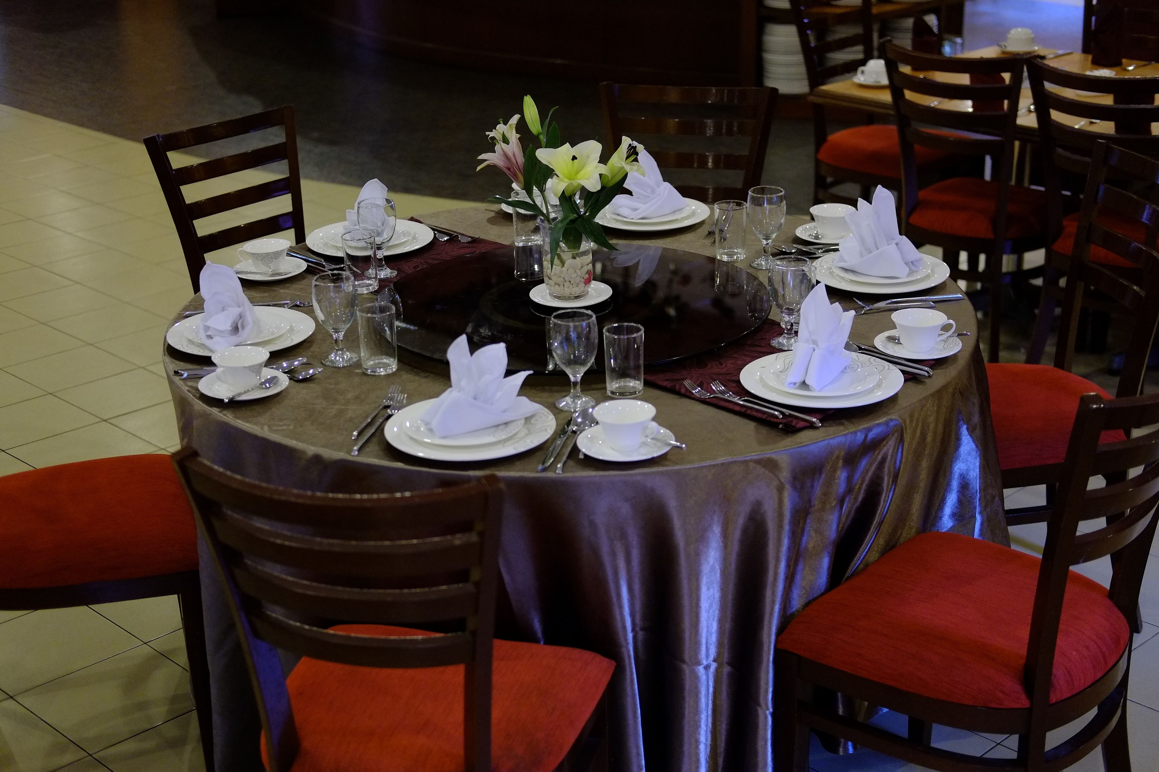 A round dining table with a dark tablecloth, white plates, and red cushioned chairs.
