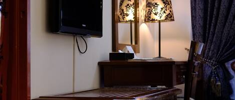 A hotel room with a flat-screen TV mounted on the wall, a wooden desk, a mirror, and a lamp with a patterned lampshade.