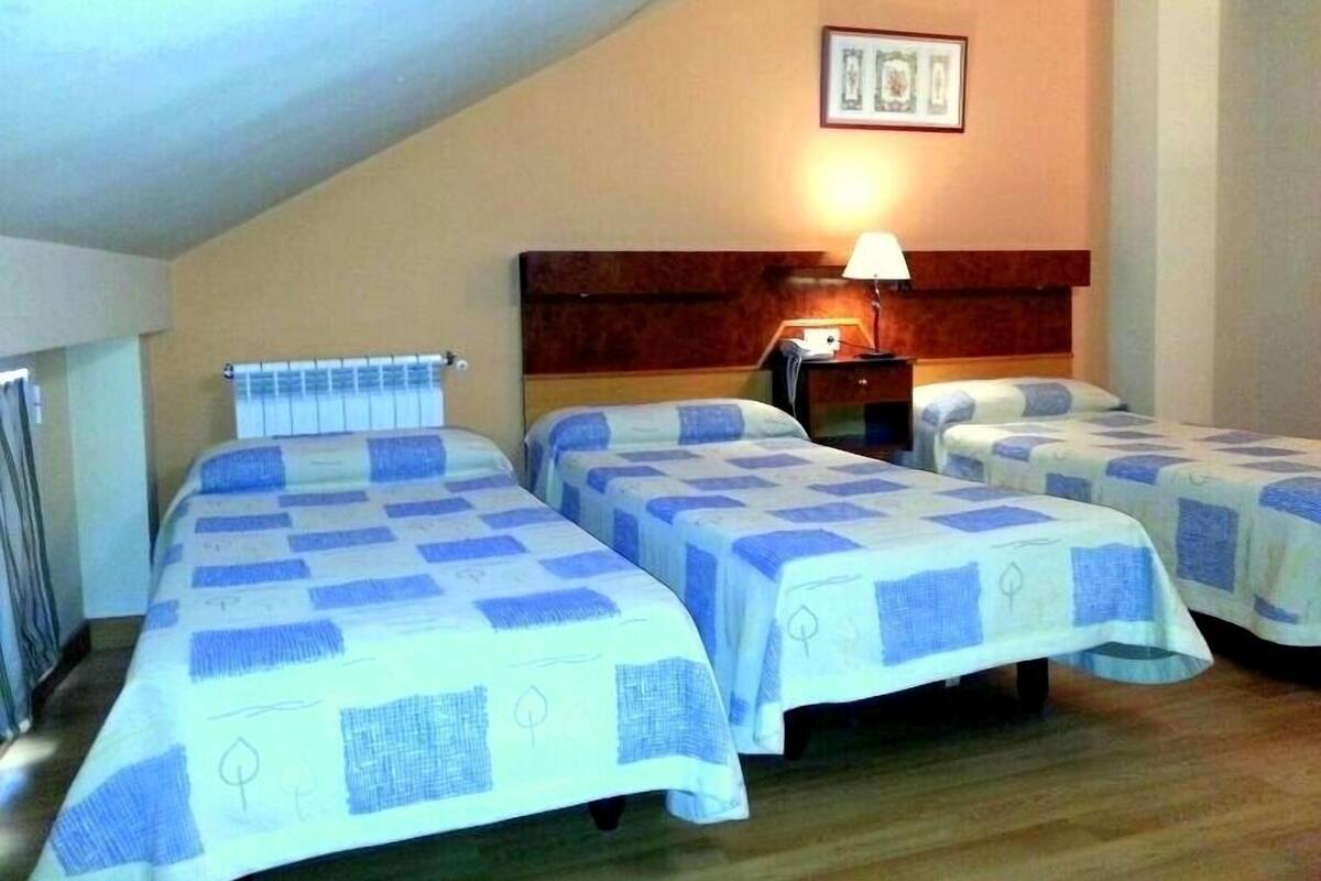 Habitación de hotel con tres camas, cada una con una colcha a cuadros azules y blancos. Se observa un cabecero de madera, una mesita de noche con una lámpara y un cuadro enmarcado en la pared.
