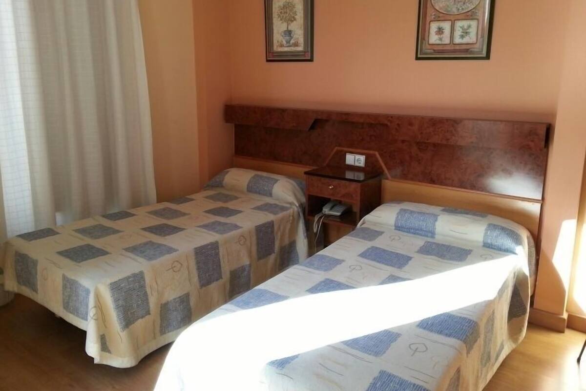 Habitación de hotel con dos camas individuales, cabecero de madera, mesita de noche con teléfono y dos cuadros enmarcados en la pared.