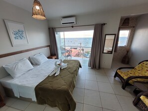 Ett hotellrum med en säng, en stol, en balkong med utsikt över havet och en väggmonterad luftkonditioneringsanläggning.