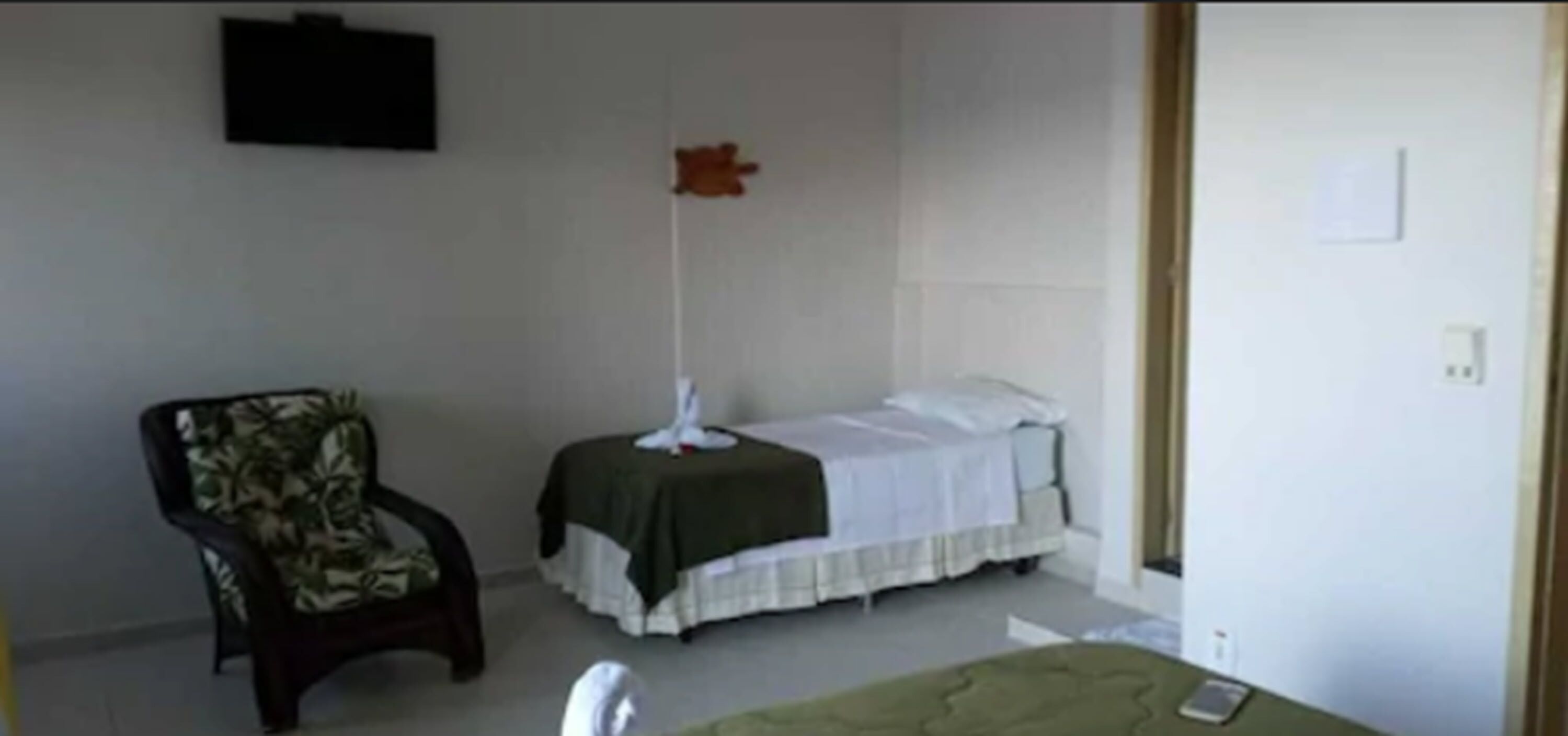 Una habitación pequeña y sencilla con cama, una silla y un ventilador de pie.