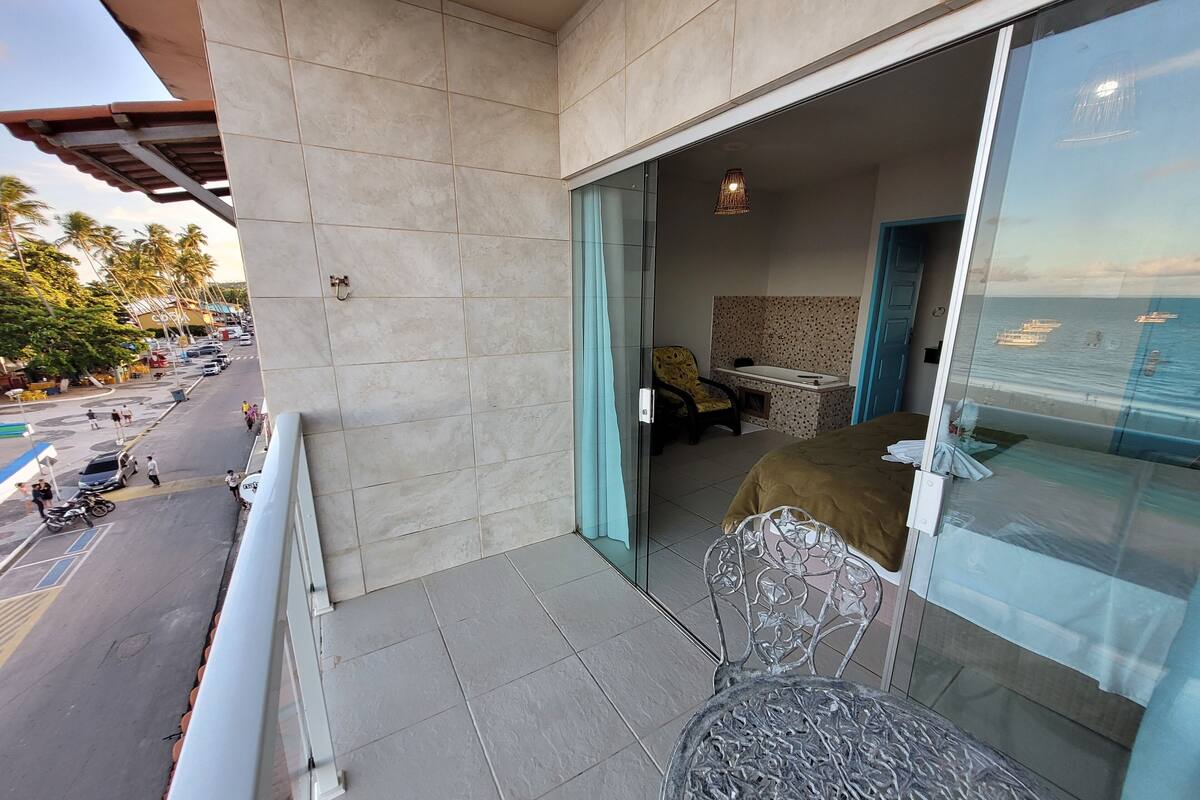 Un balcón con vista a la calle y al océano. Se ve una cama con vista al océano, una silla y una mesa.