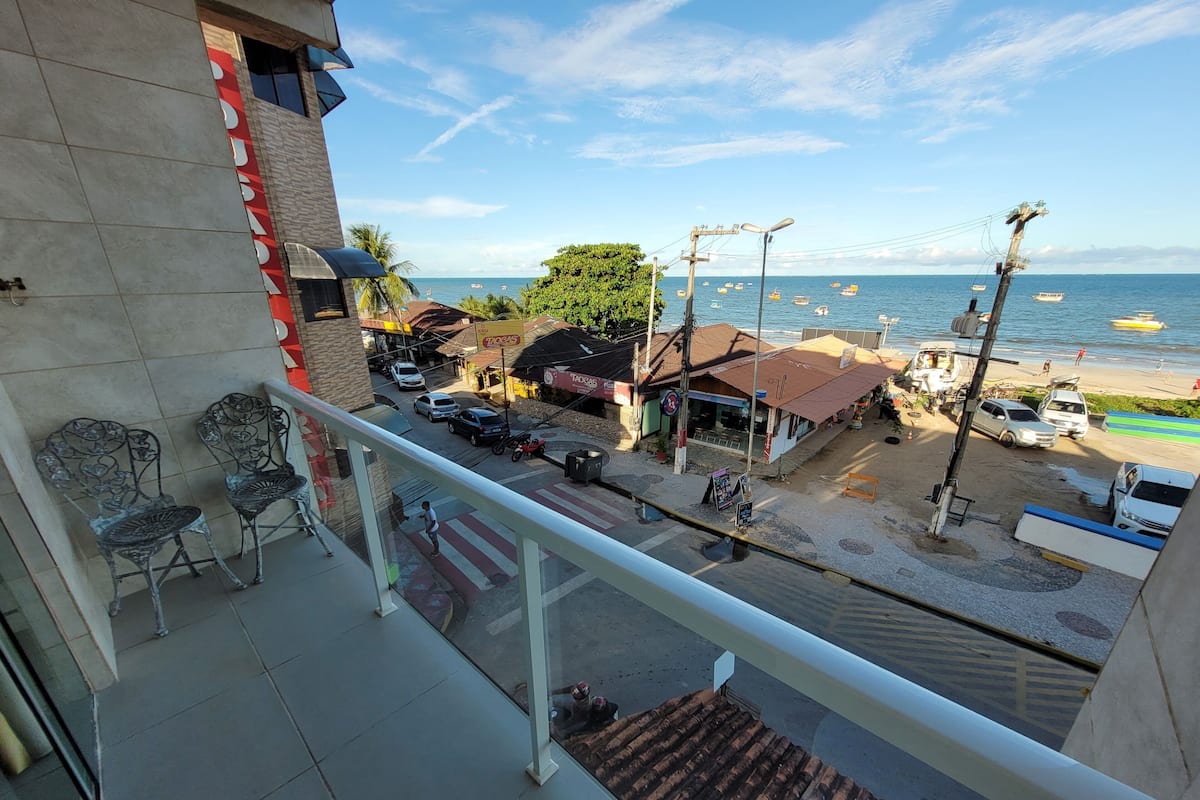 Un balcón con sillas de metal, vista a una playa con barcos y una calle con tiendas y coches aparcados.