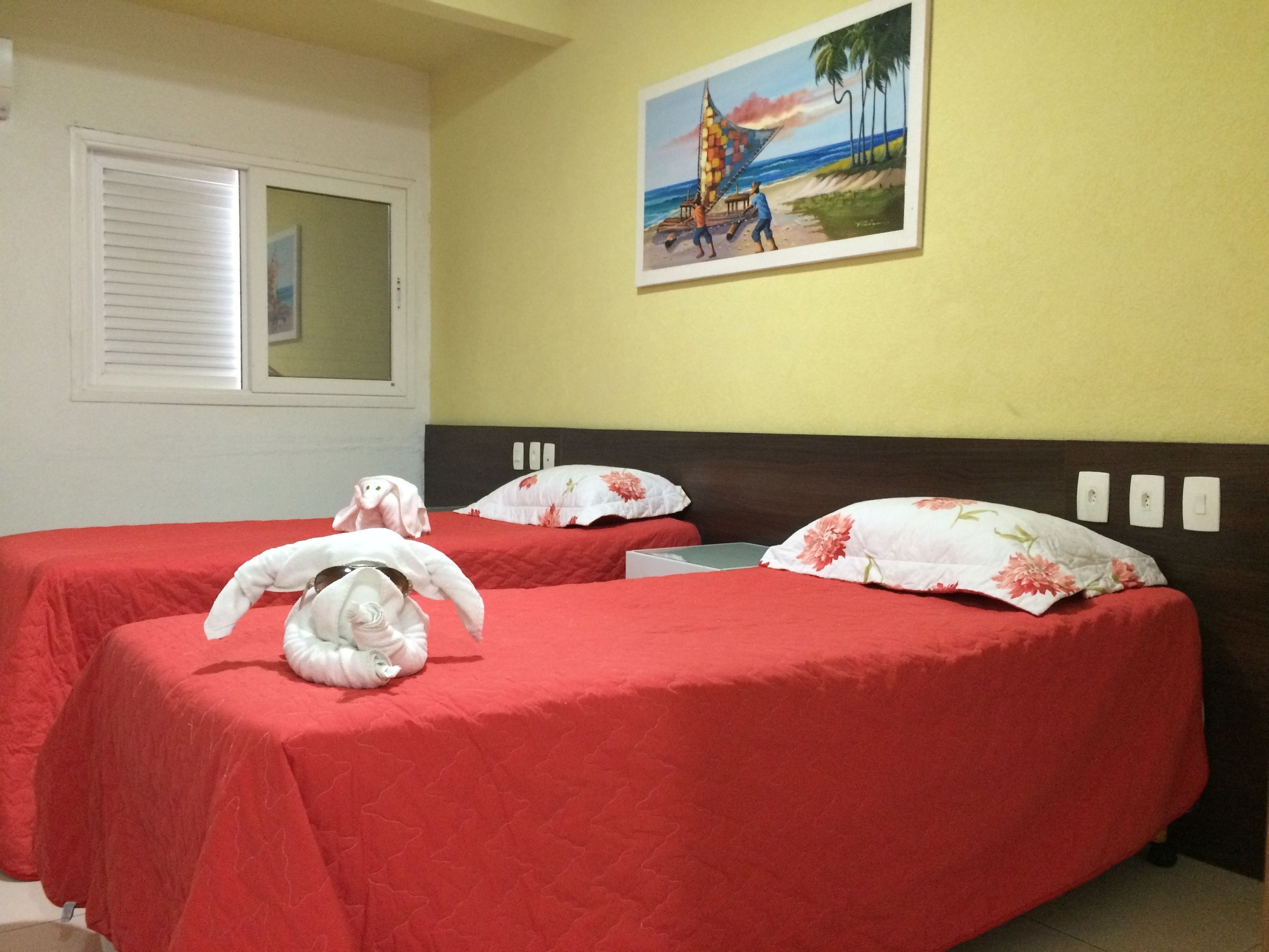Ein Hotelzimmer mit zwei Betten, beide mit roter Bettwäsche und floralen Kissen. An der Wand hängt ein Gemälde einer Strandansicht.