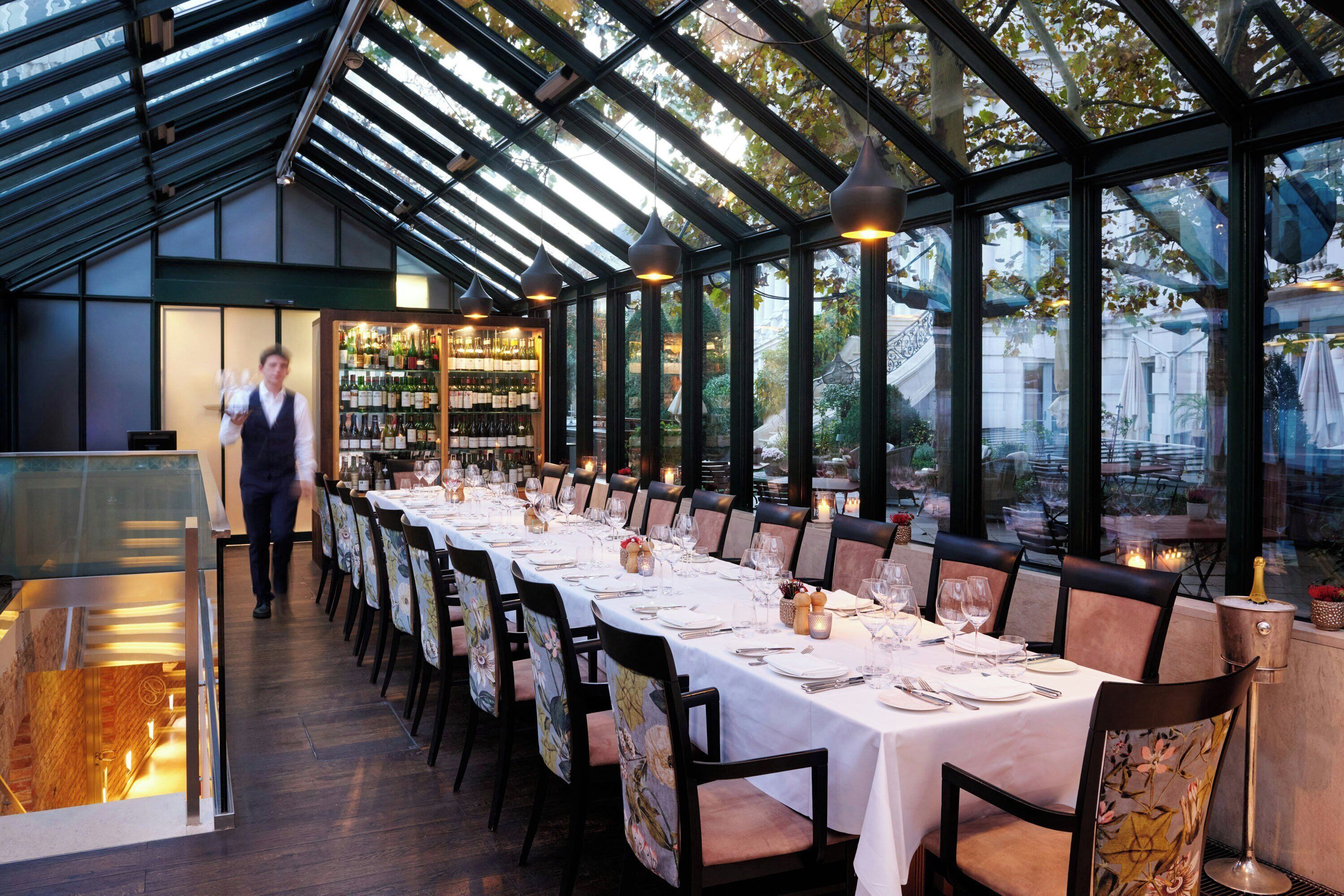 A restaurant with a long table set for dining, a wine rack in the background, and a view of trees through large windows.