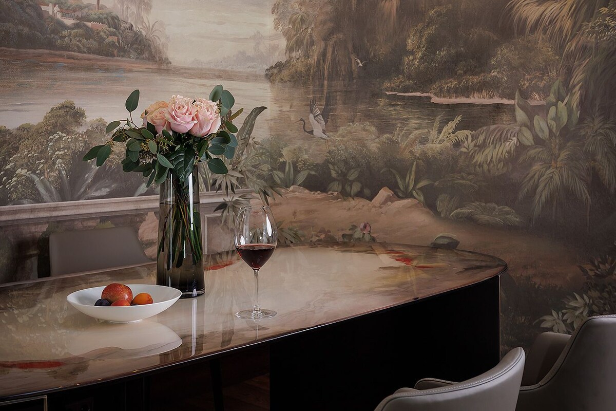 A dining area with a table set for two, a vase with flowers, and a painting of a landscape on the wall.