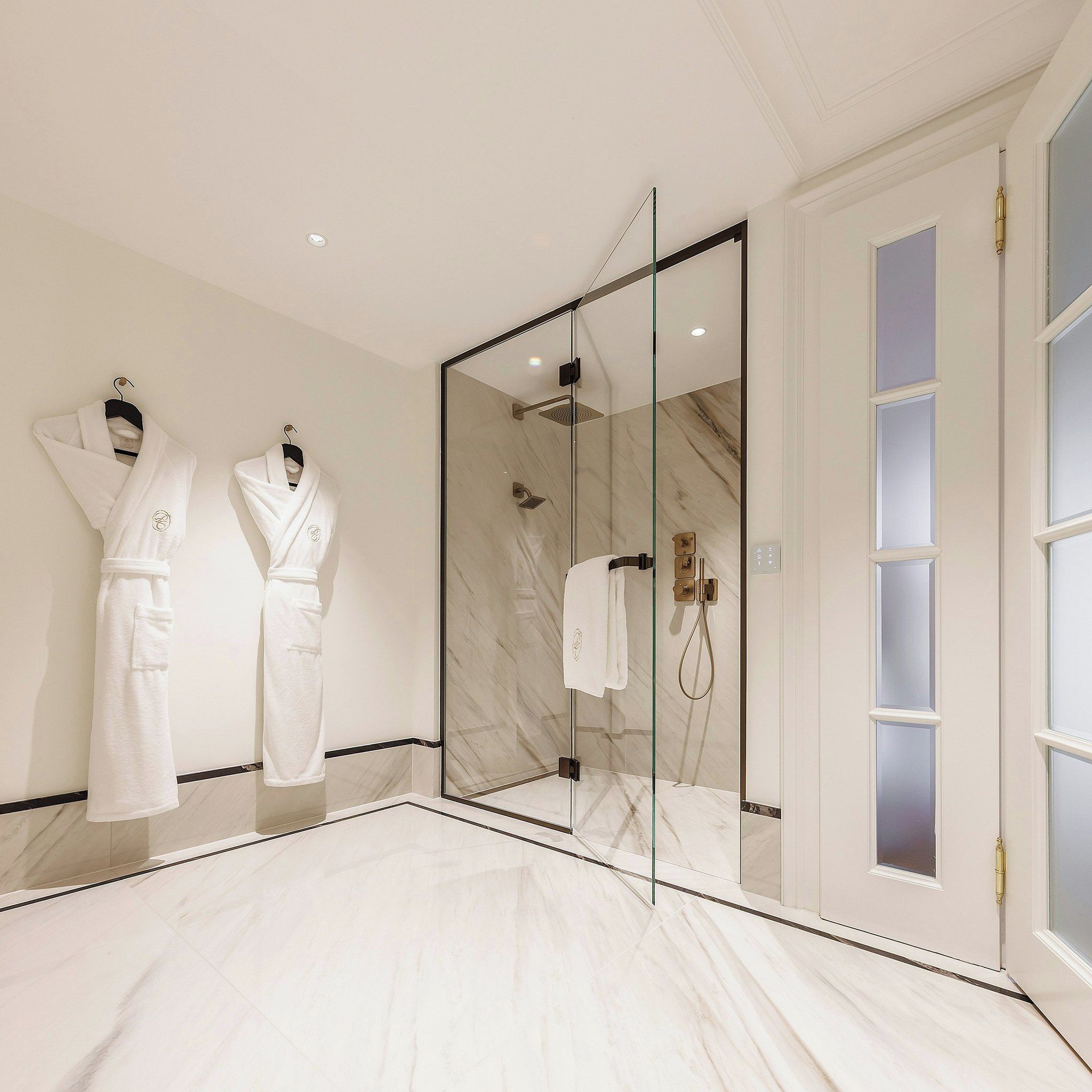 Un baño moderno con ducha envuelta en vidrio, suelo de mármol y dos albornoces blancos colgados en la pared.