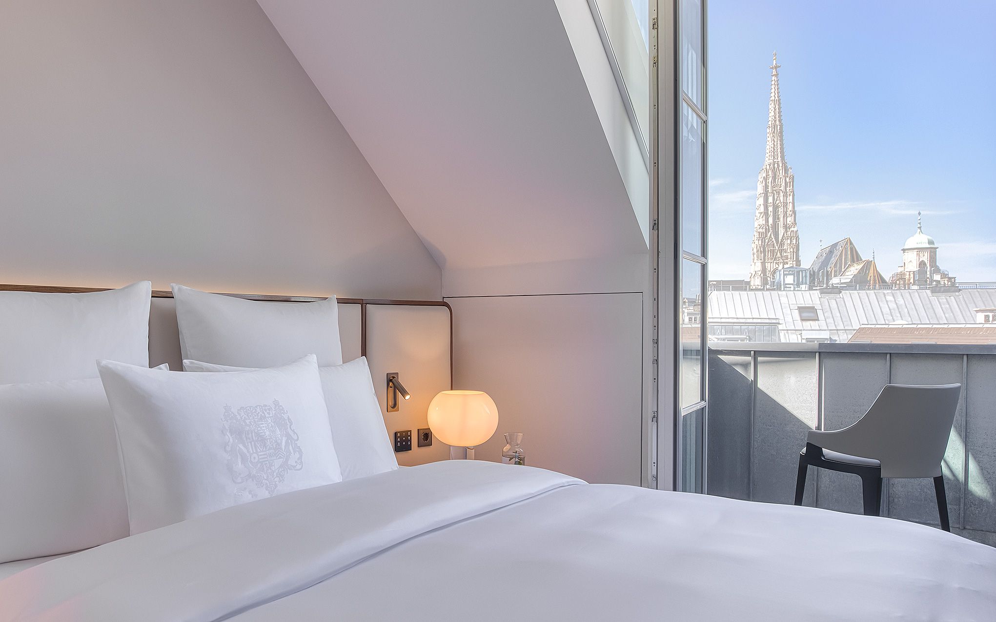 A hotel room with a bed, a nightstand, a lamp, and a view of a cathedral.