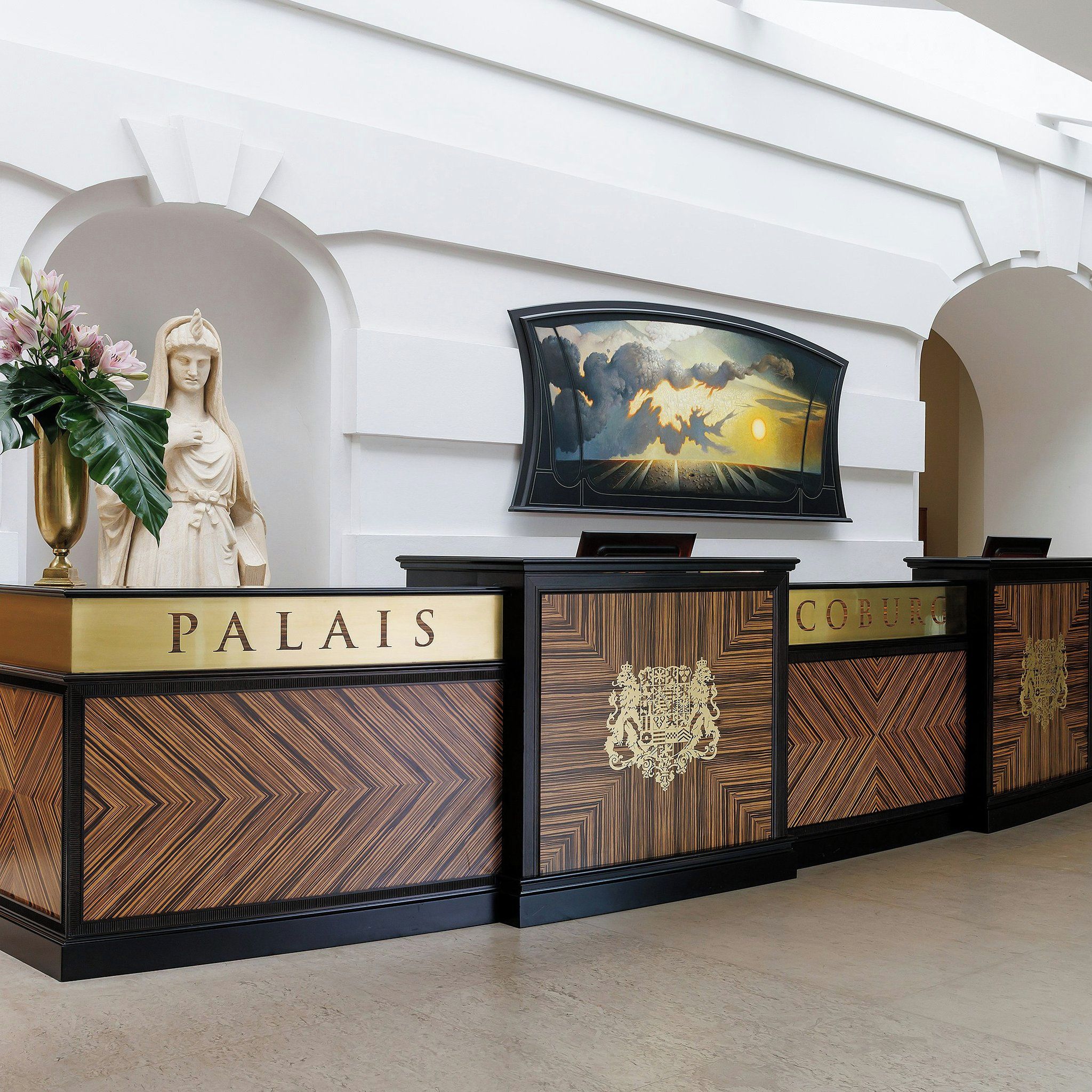 A reception desk with 'PALAIS' and 'COBURG' signage, a statue, a television screen, and a decorative wall.