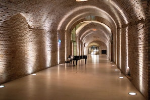 A long, arched brick corridor with a grand piano and a bench, illuminated by recessed lighting. - Palais Coburg  (Vienna)